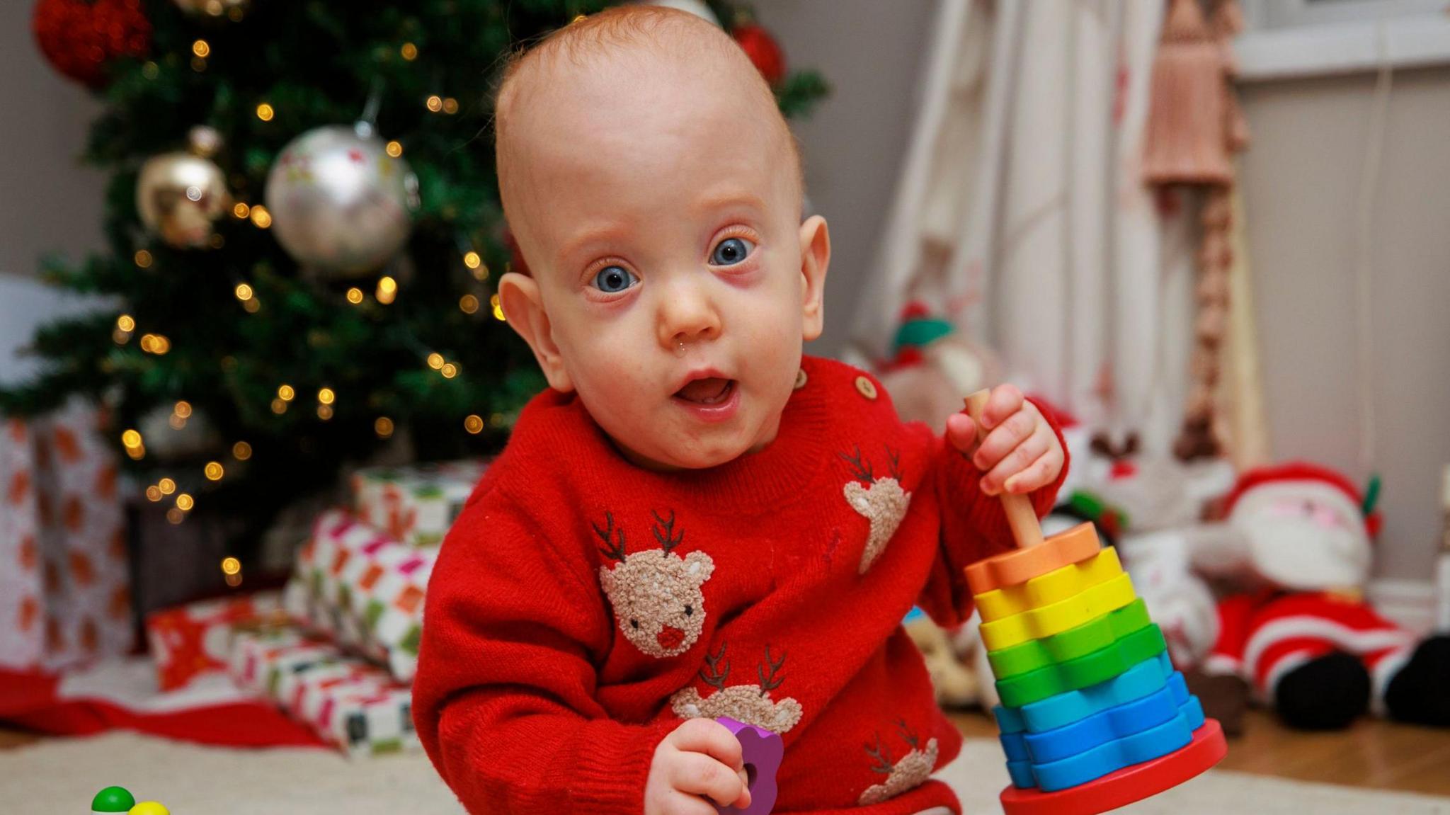 Iarla Ace sits on a cream carpet playing with toys. He has a red jumper with Rudolph the reindeer on it. Behind him is a Christmas tree and wrapped presents.