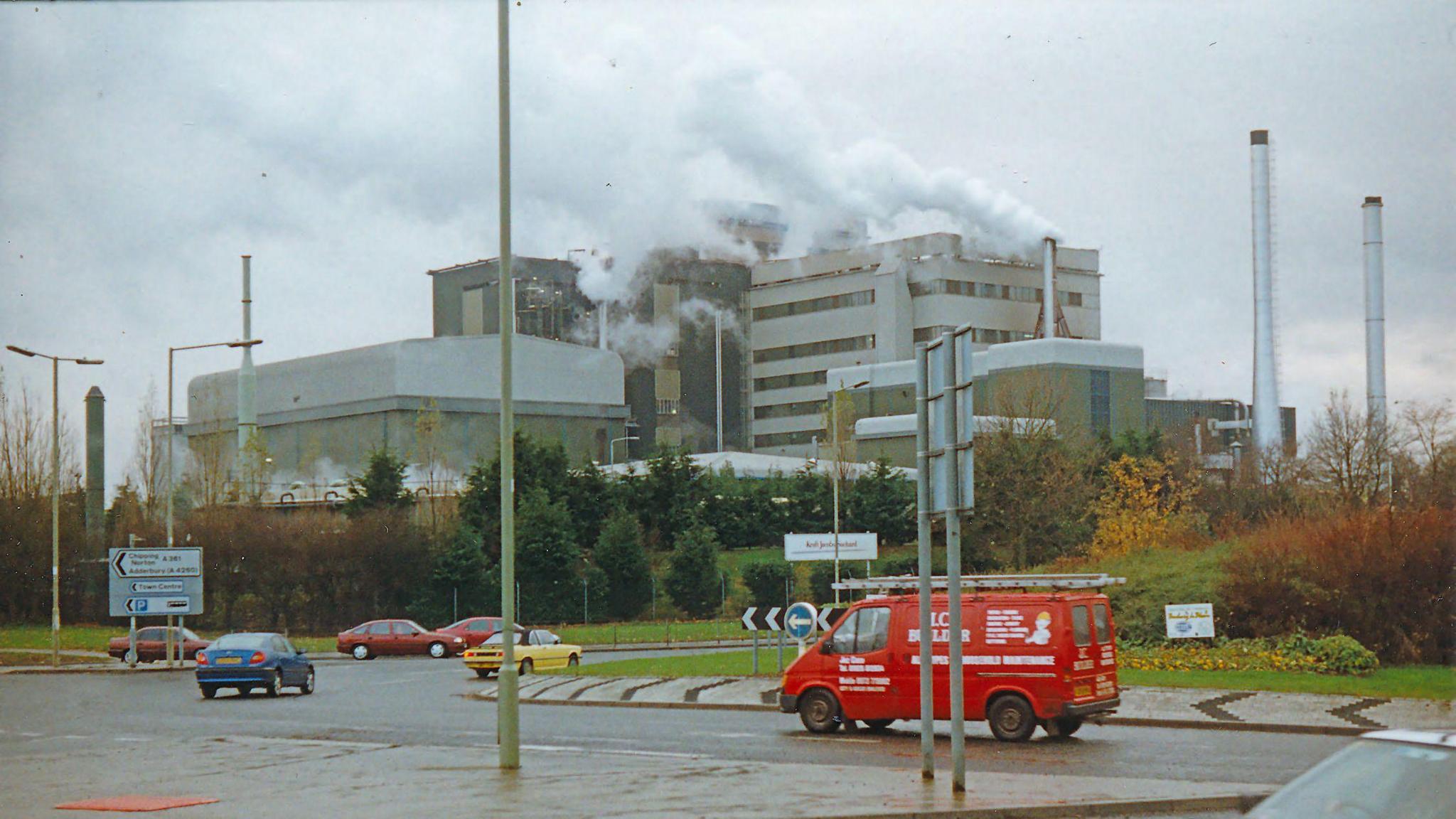 The large grey and green structure rises above a roundabout as large plumes of smoke are emitted from its large chimneys.