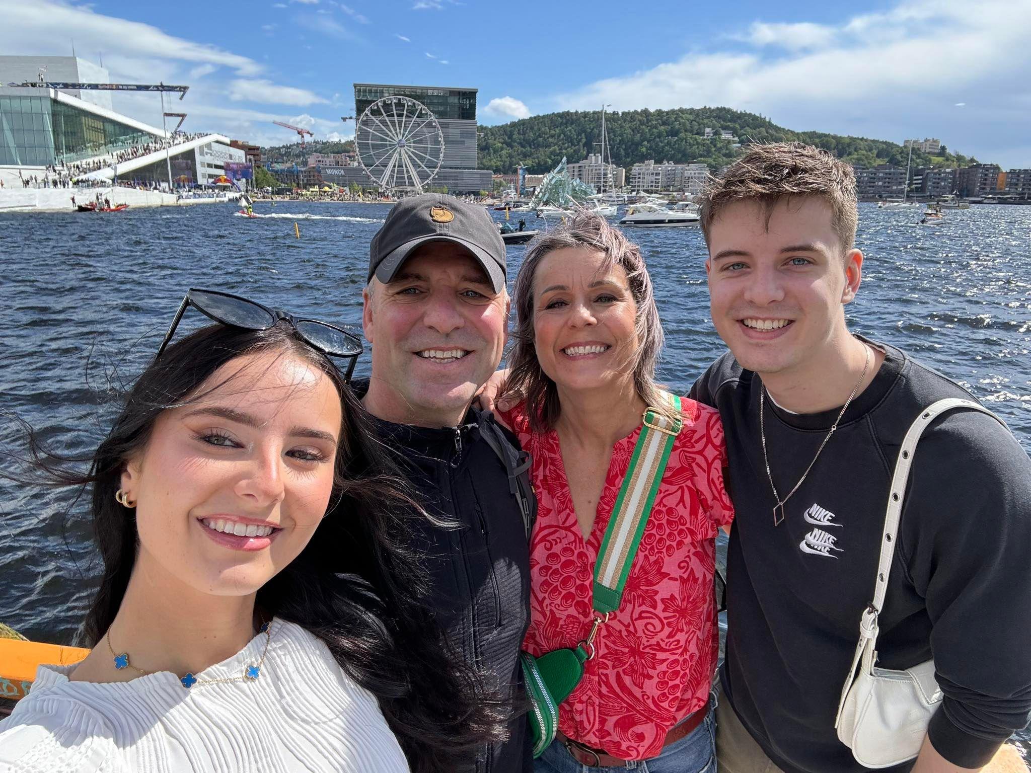 Jeannie Ambrose, her partner and two children are stood in front of a marina. They are all smiling at the camera. In the background there are boats and a Ferris wheel