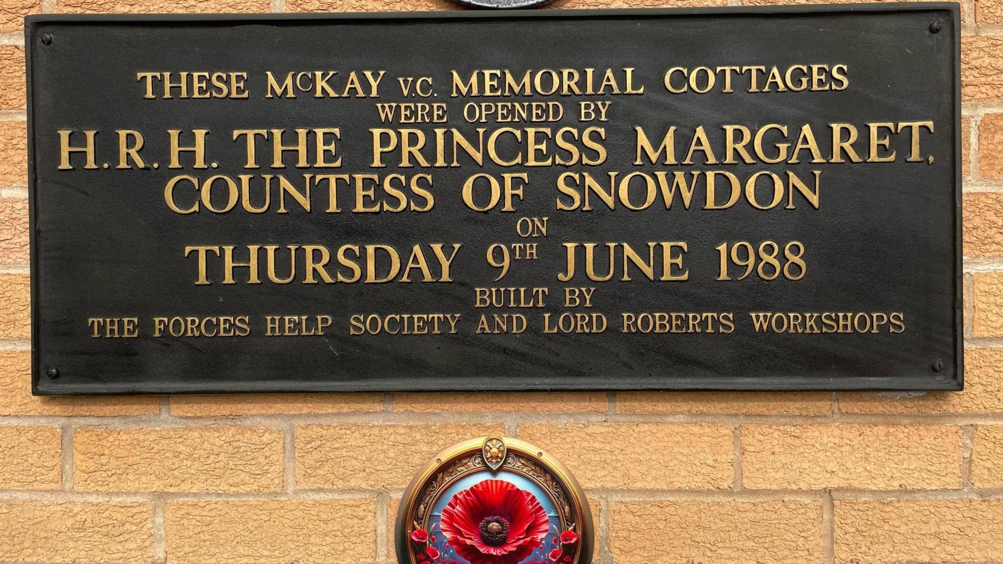 A plaque on the wall of a house with a poppy underneath