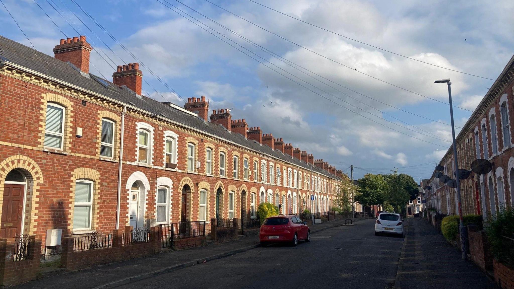 A street with red-brick houses on either side. The houses are attached, and there is a car parked on either side of the street. The weather is sunny.