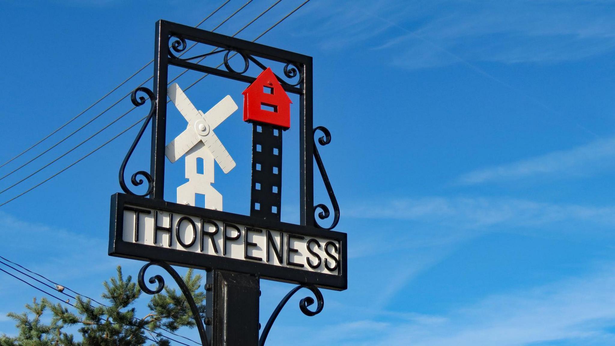 Metal village sign that says "Thorpeness", made from metal and featuring representations of a windmill and The House in the Clouds, a water tower disguised as a house.