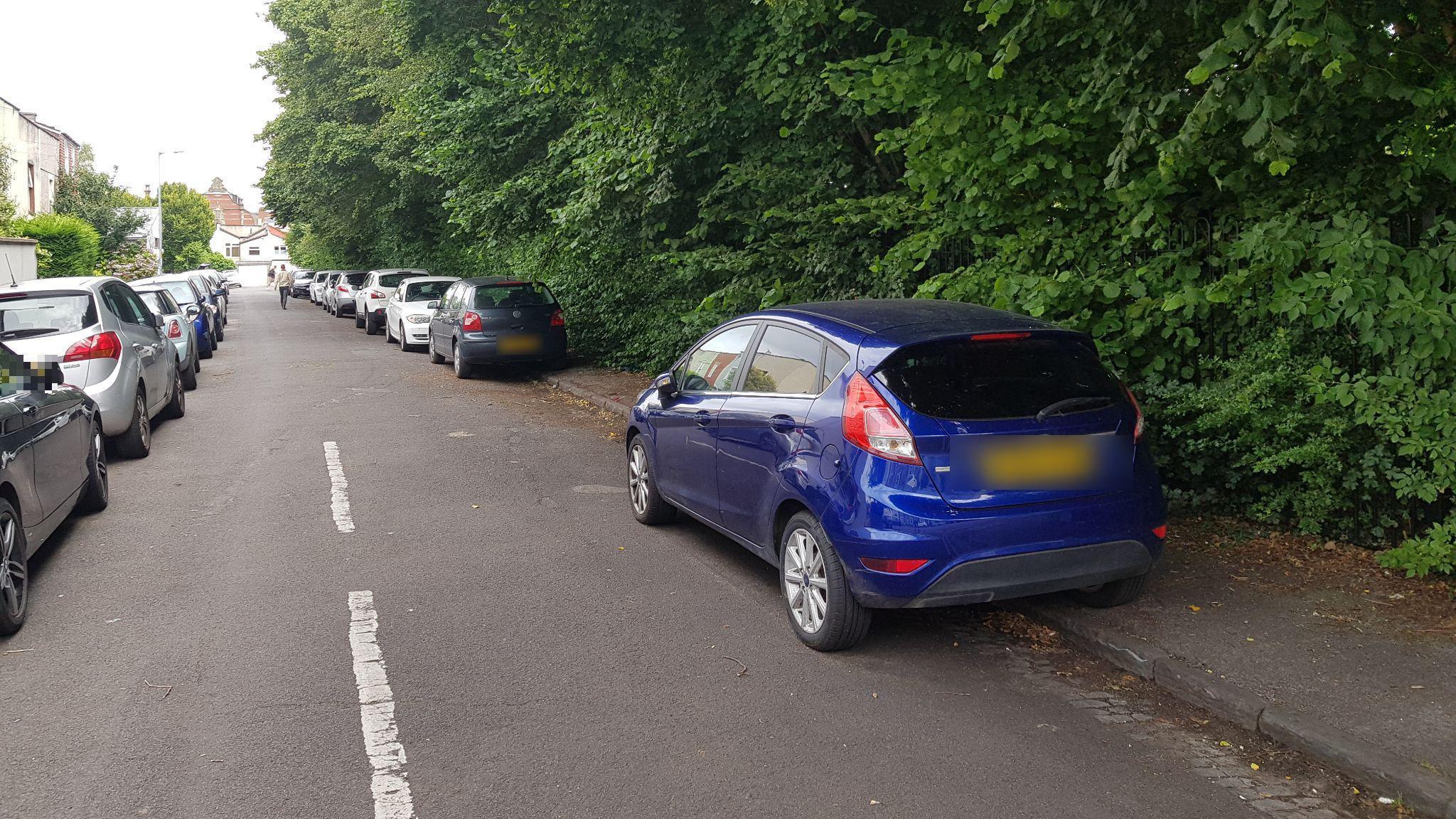 A residential street with cars on the right hand side partly parked on the pavement