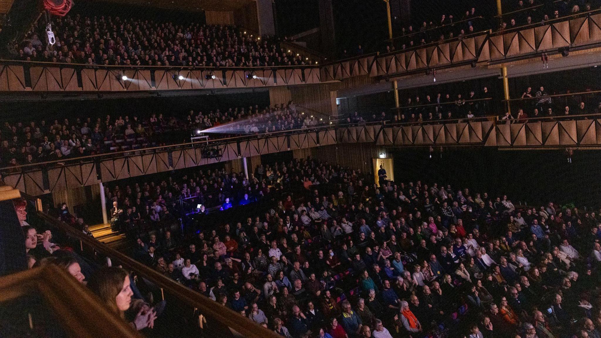 A packed-out Bristol Beacon main hall is seen full of audience members watching the gala evening at the Slapstick Festival. The auditorium is largely in darkness with a projector beam coming from high up at the back.