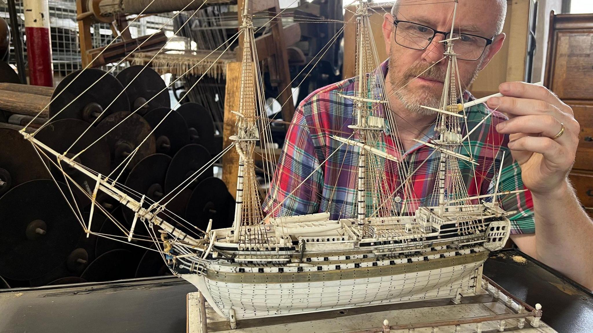 A man cleans a model ship made of ivory using a tiny paint brush
