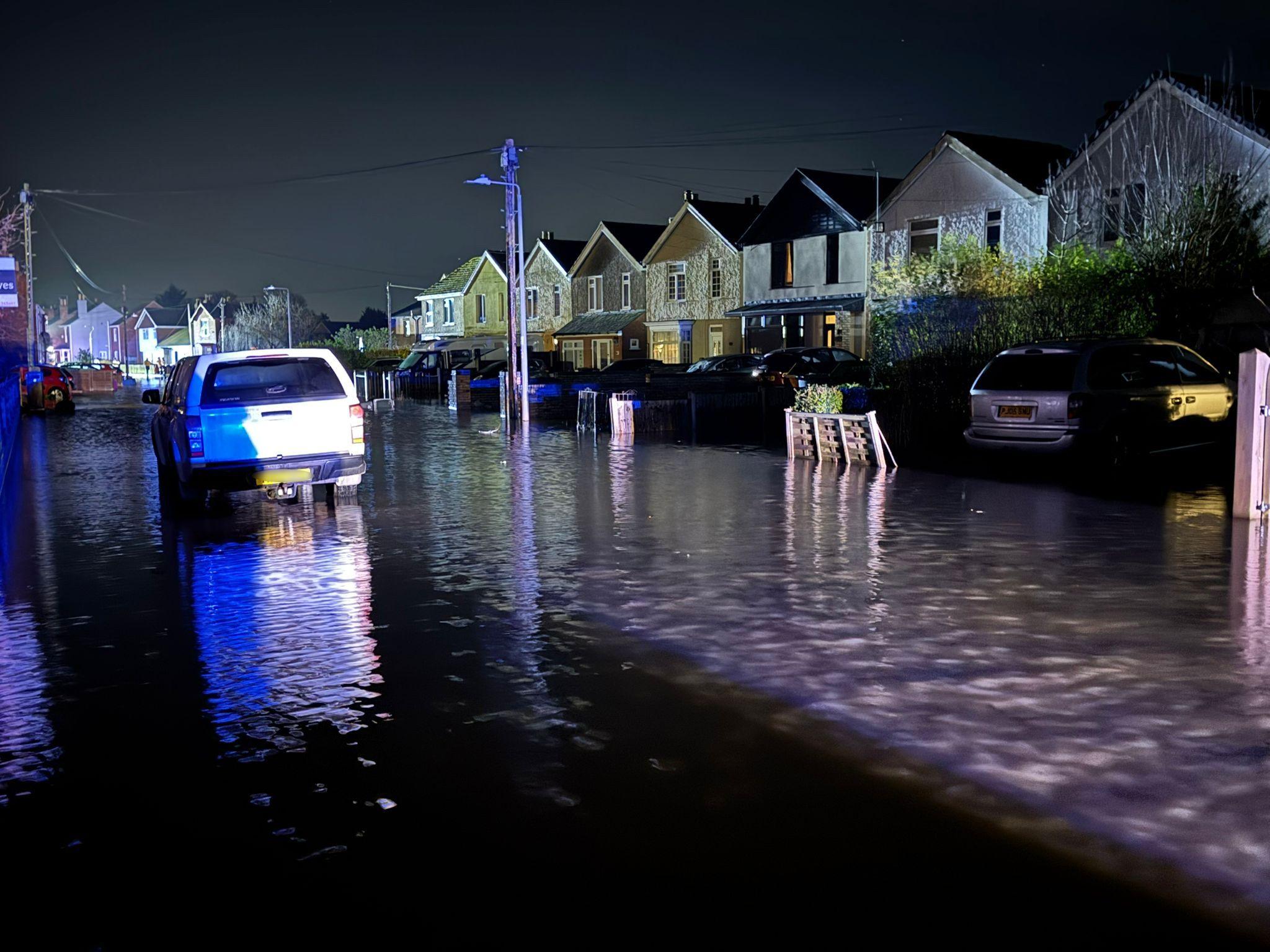 A flooded residential street at night. In the distance is a row of houses with cars parked on the drives. In front is water flowing down the street with a pick-up van parked on the right. The flood water is nearly reaching its rear bumper