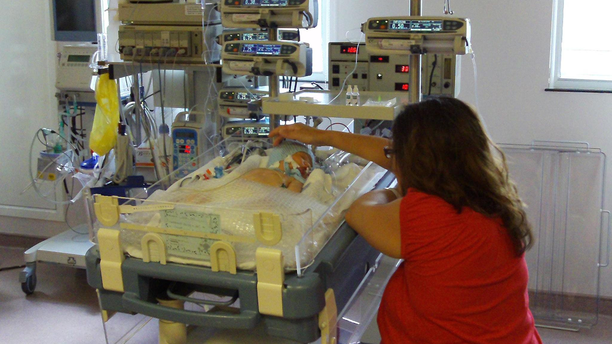 A mother sat next to her baby in an intensive care unit cot. She has long dark hair and is wearing a red T-shirt. Her back is to the camera and she has one hand stretched out to touch her baby boy's head. The baby is lying in the cot with a white blanket over him. Medical equipment is around him.