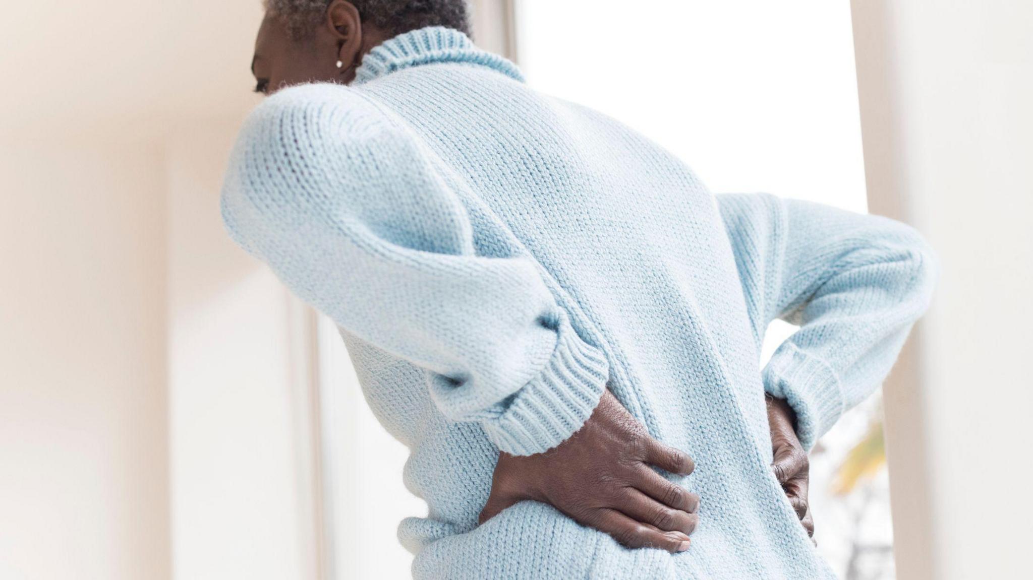 A woman wearing a pale blue jumper is seen from the back. She is leaning forward and has her hands placed on her lower back.