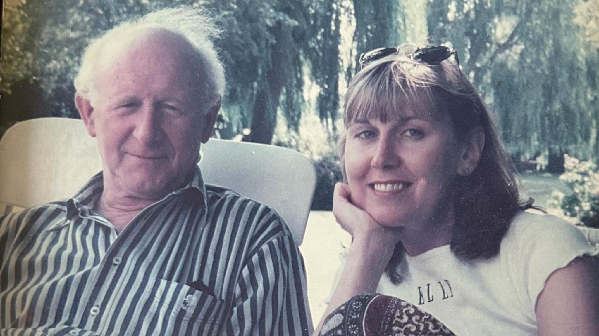 An old photograph with faded colours of Jane Arnold smiling with her father Barry on a sunny day in a garden. Jane has blonde hair and has sunglasses on her head. Barry has white hair and a striped shirt.