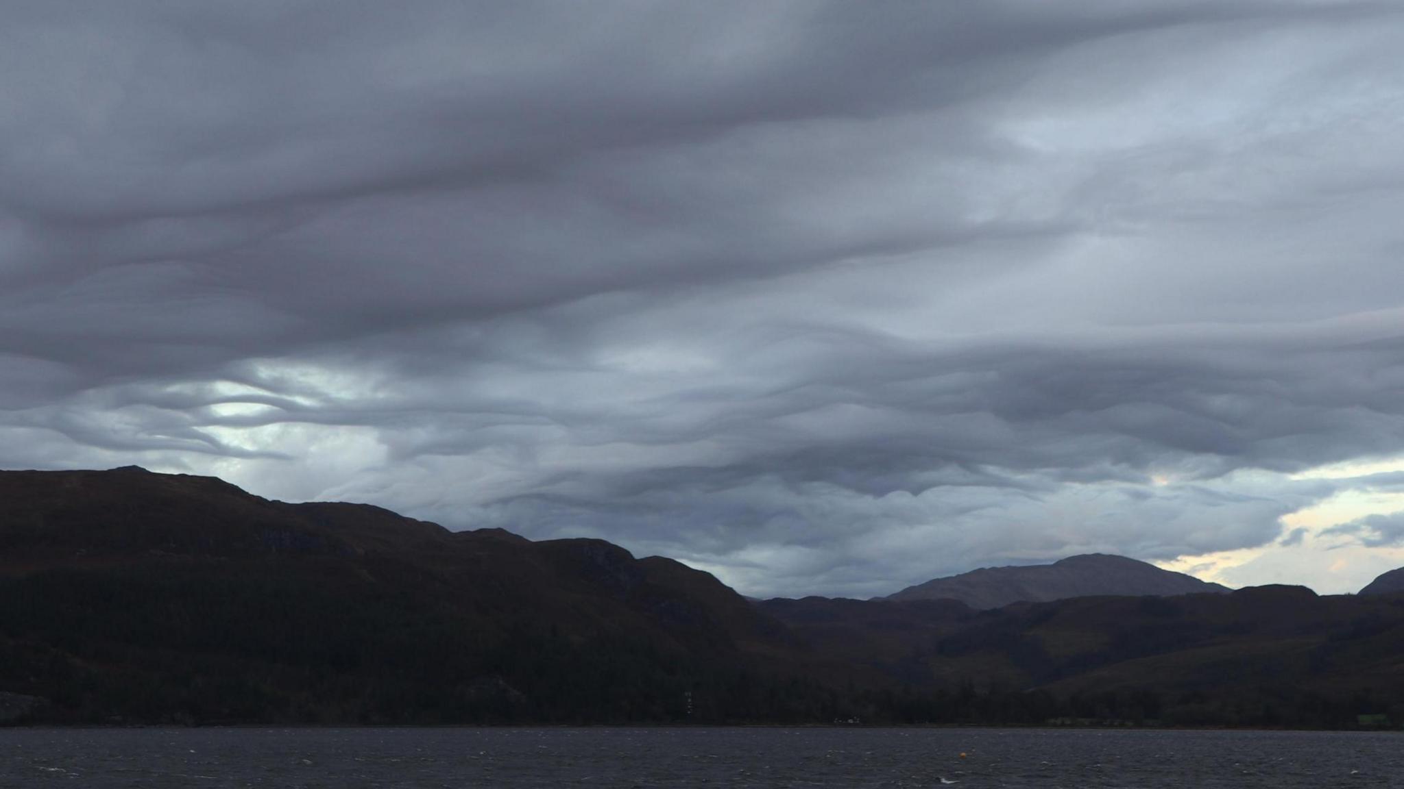 Wavy dark grey clouds appear in the sky over mountains and a loch in the Highlands.