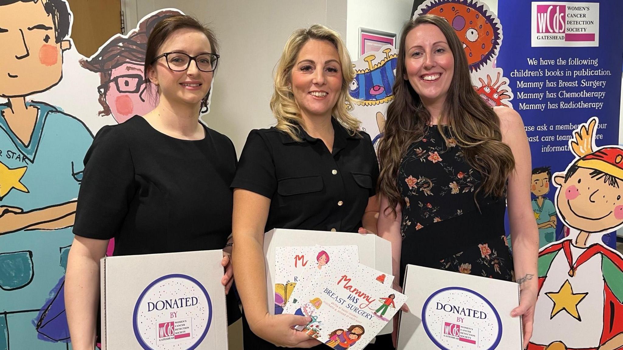 Rachel Lockerbie, Kathryn Jobes and Emily Turnbull are standing in a line and smiling brightly at the camera while holding up the children's books and the boxes they come in. Stickers on the boxes read: "Donated by Women's Cancer Detection Society".