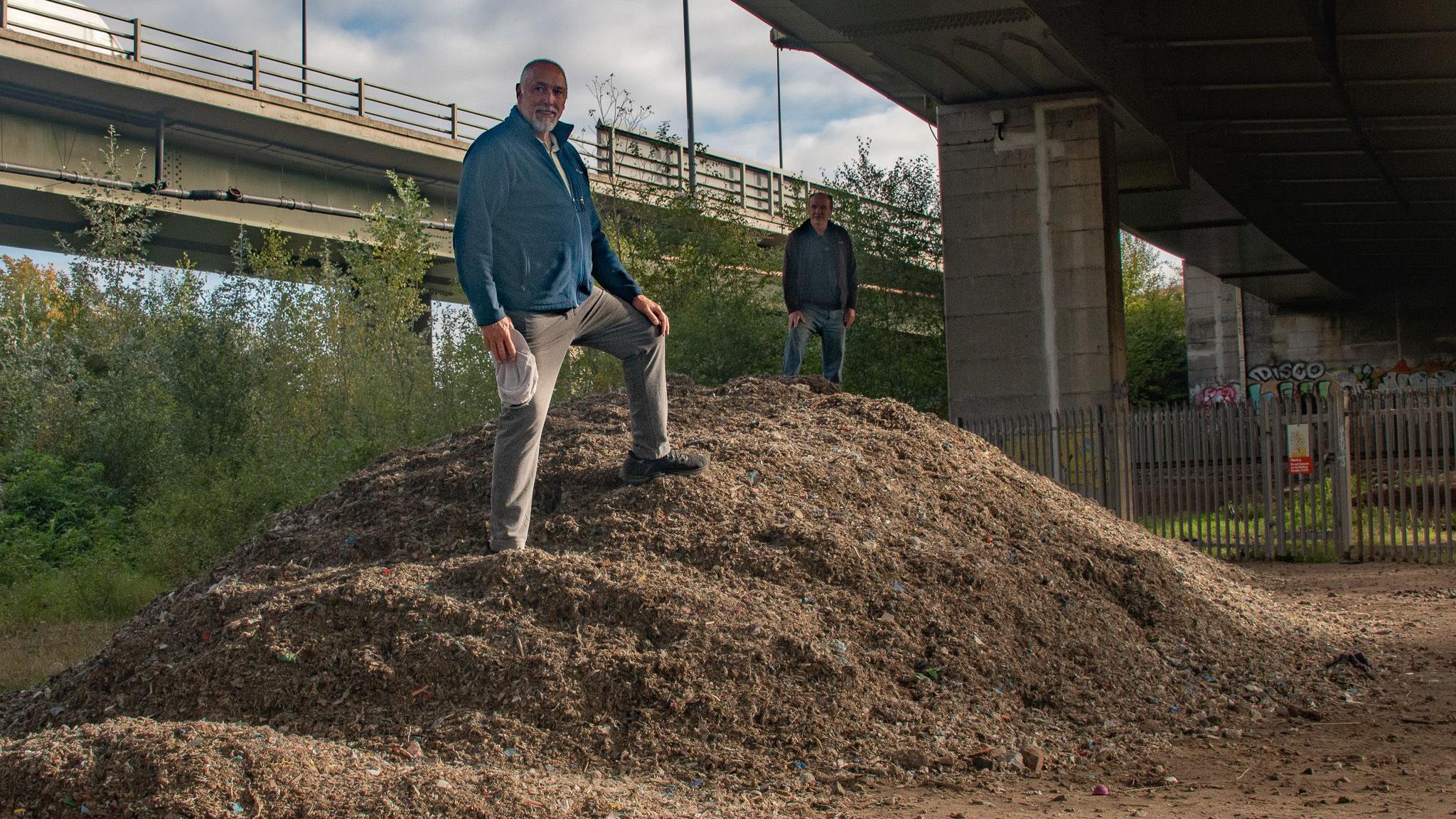 Steve Taylor, who is bald and has a white goatee beard, and Alan Howgate wearing a burgundy fleece, stand on a mound of rubbish beneath a motorway bridge.