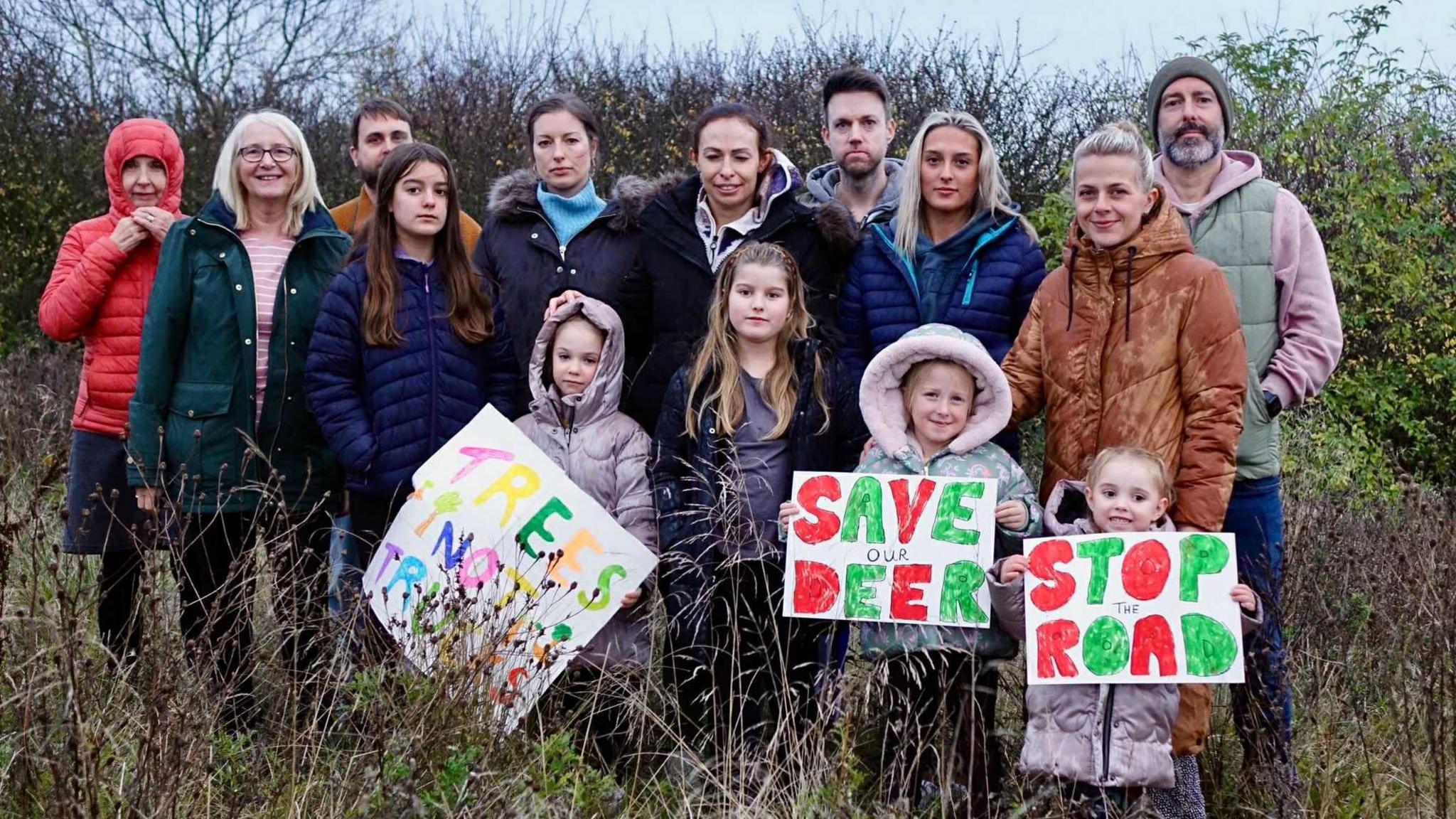 A group of 14 people opposing the plans standing in a field. Three of the four children standing in front of the group of adults are holding signs reading Trees Not Trucks, Save Our Deer and Stop The Road.
