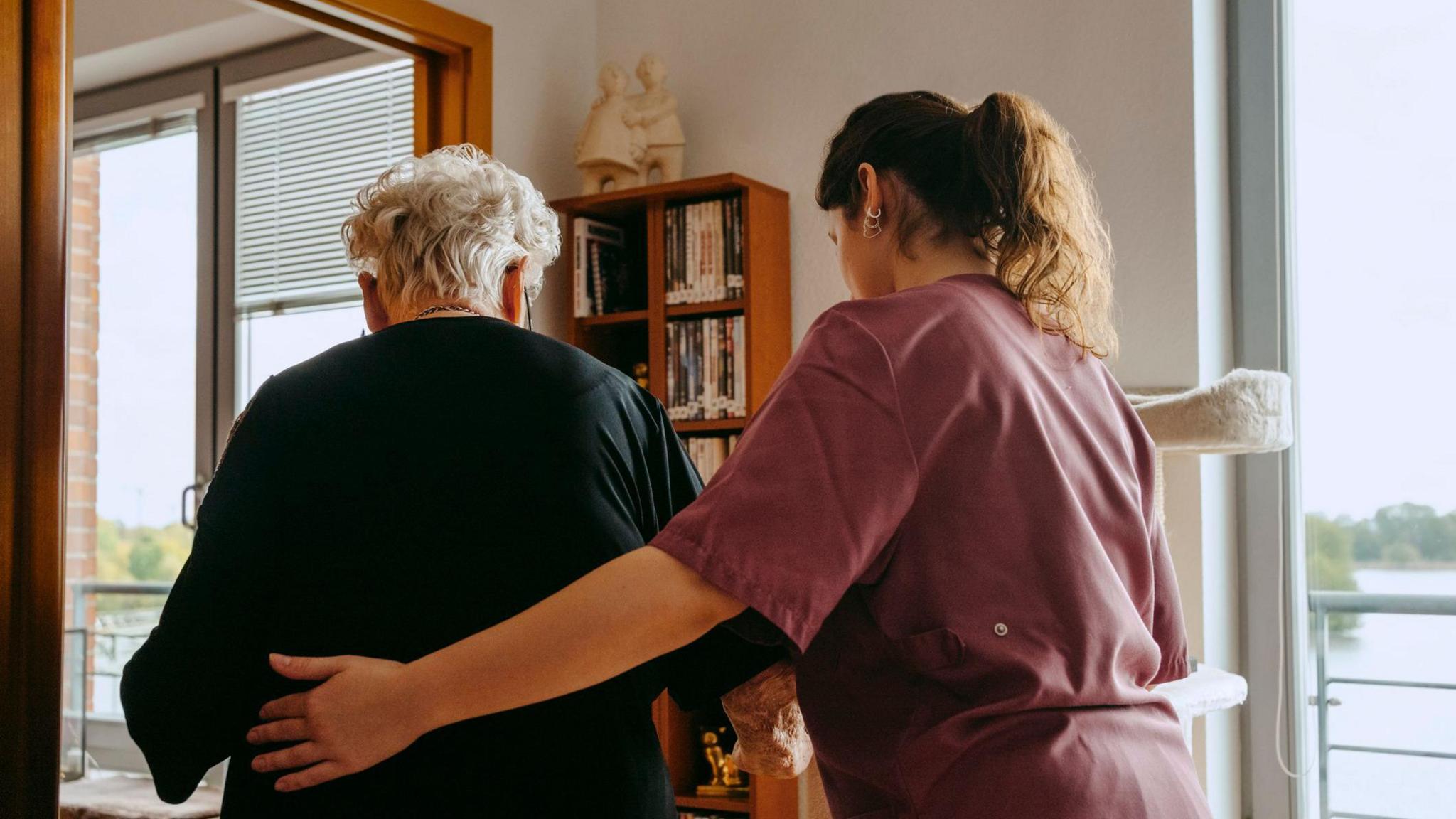 An elderly woman with short white hair is escorted by a woman with long brown, wearing maroon scrubs. The younger of the two has her left hand on the woman's back. The elderly woman wears a black dress. A wooden book shelf, two shelves wide and four long is partially stacked with books. There are two windows either side of the bookshelf, a body of water is visible outside.