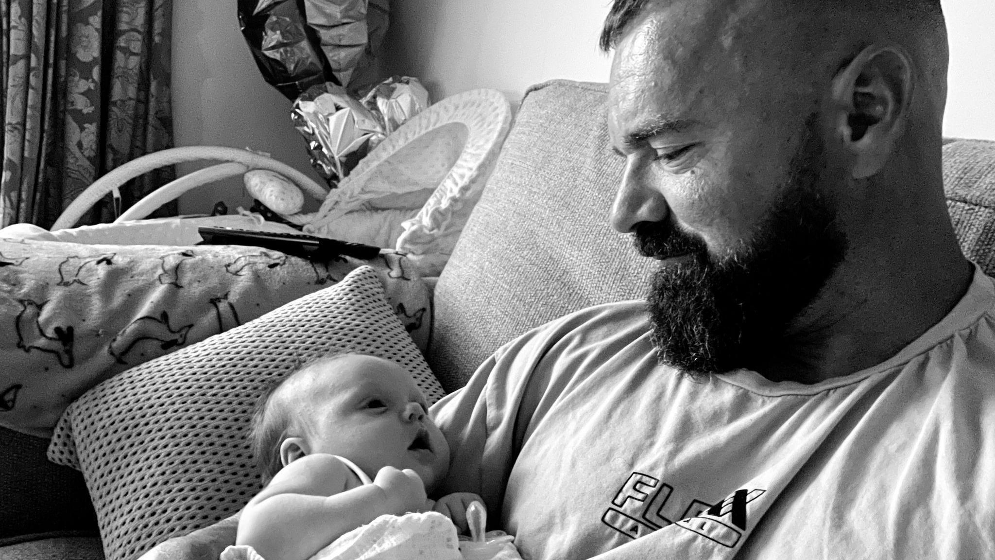 Llun du a gwyn o Mat yn eistedd ar soffa, yn edrych yn serchus ar ei ferch fach yn fabi yn ei freichiau