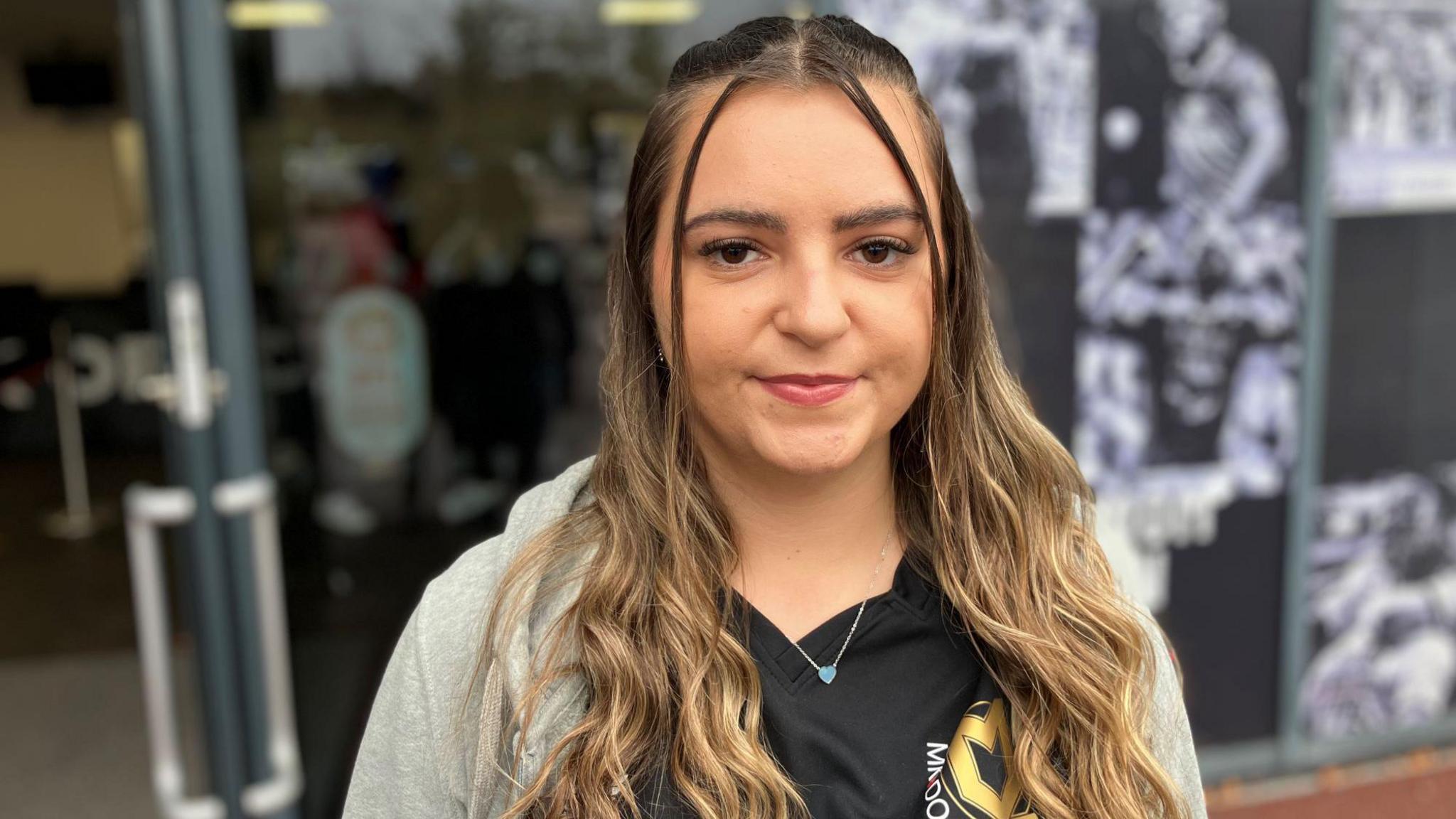 Young woman with long blond and brown hair wears a MK Dons football shirt and stands outside the club entrance.