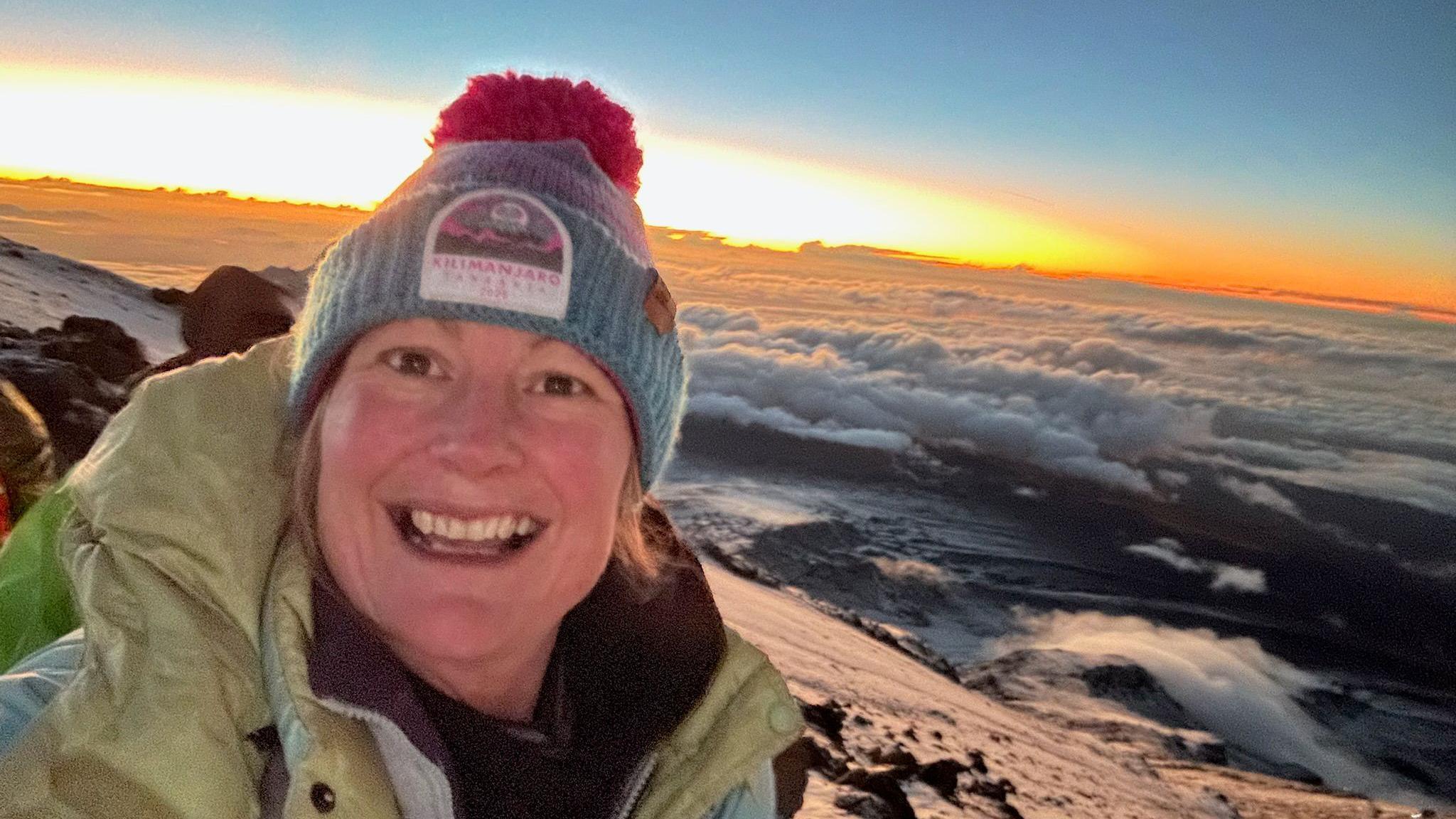 Gill, who is wearing a blue and purple hat with a purple pom pom, takes a selfie with the slopes of a mountain and a sun rise behind her.