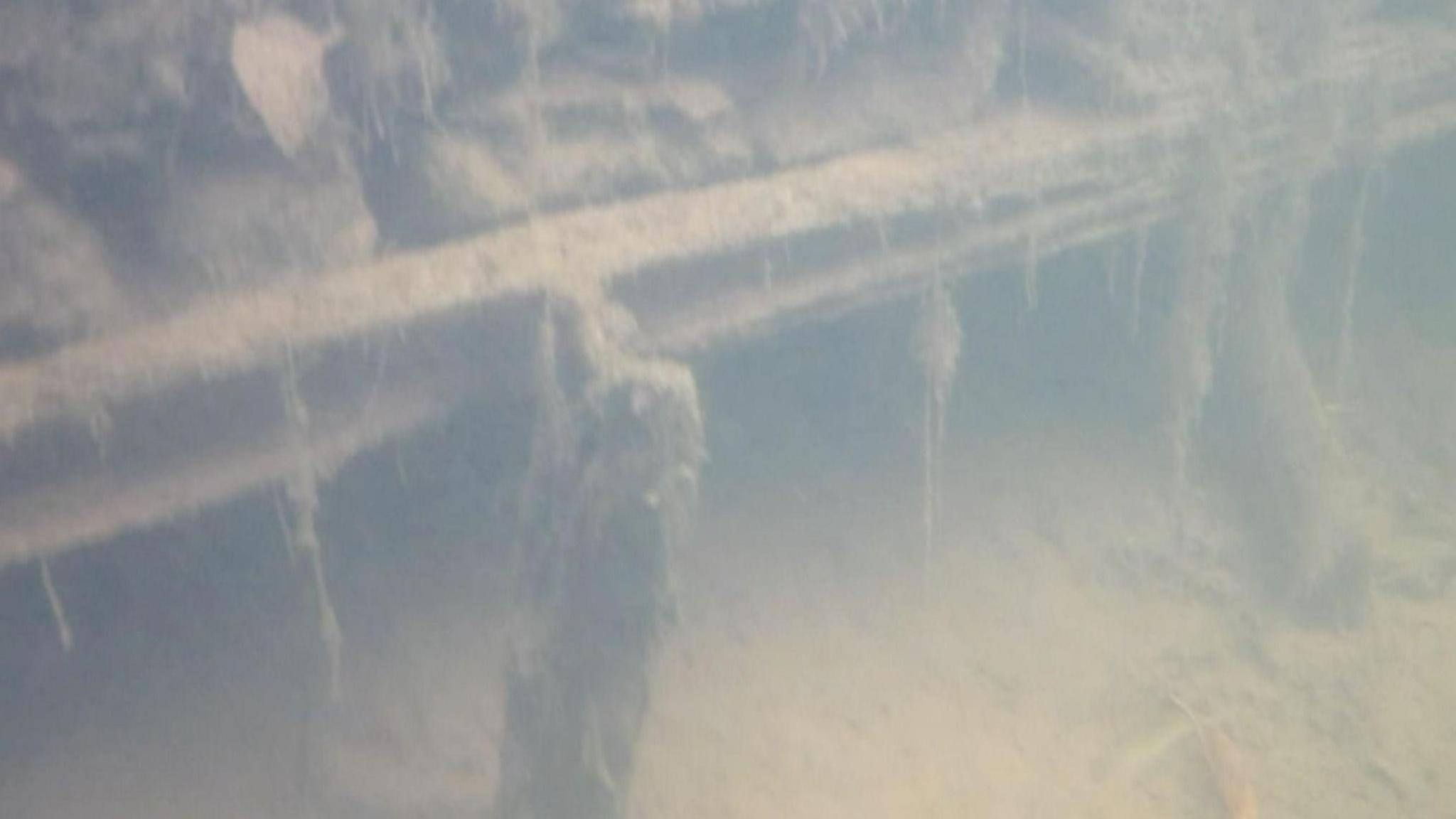 A grainy grey image of a bridge structure under water. The metal structure, which has dirt and plants stuck to it, looks worn and part of it has given way.