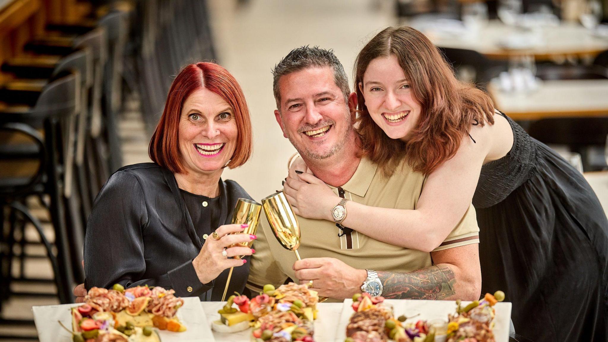 Ian Steele smiles and laughs as he sits next to his his wife, Nick who has short red hair and is wearing a black silk top. Ian has short dark hair and is wearing a cream coloured polo top. Chloe embraces her dad around the shoulders, wearing black dress and has long brown hair, she is smiling at the camera.
