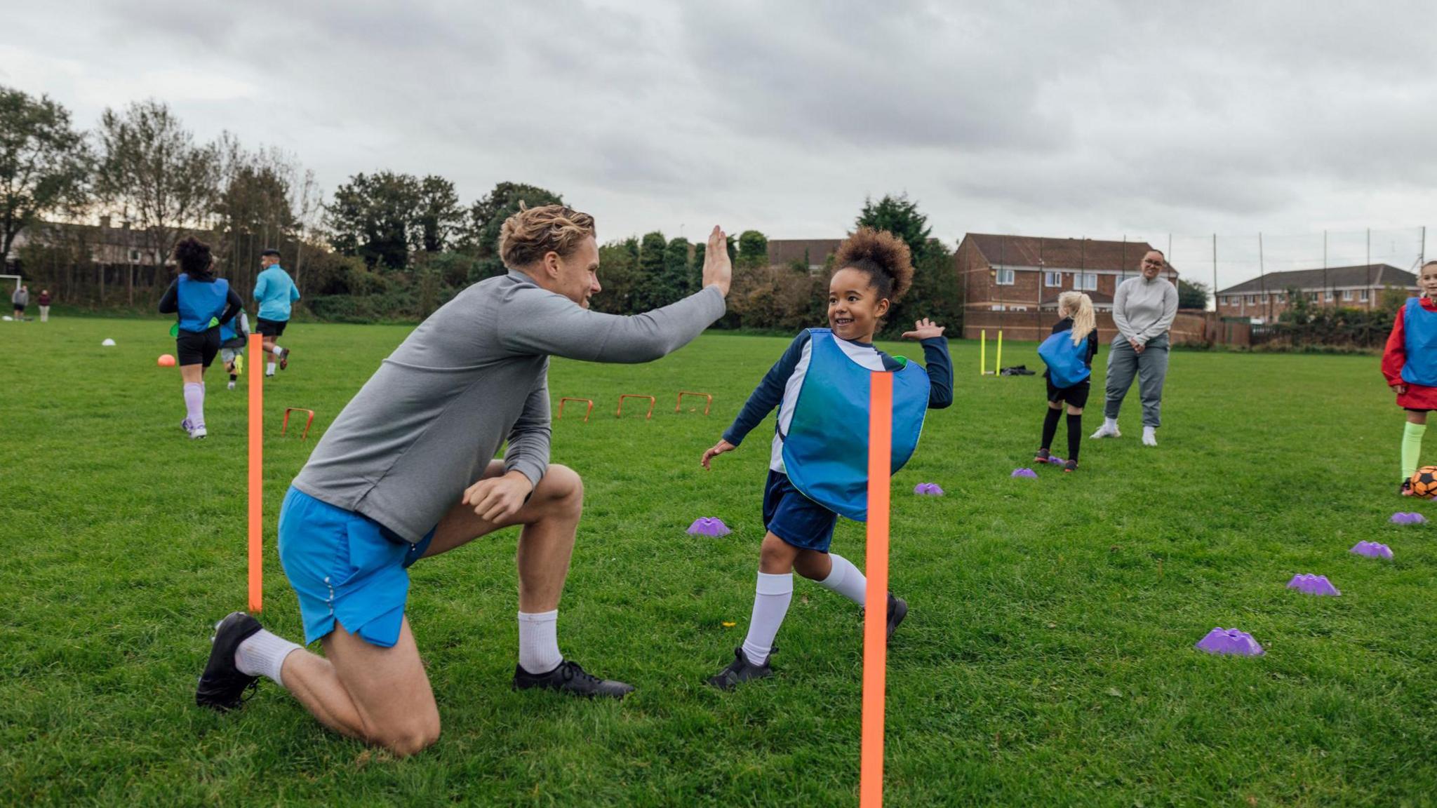 A wide shot of football coaches and a group of children wearing sports clothing, football boots and sports bibs on a football pitch in the North East of England. A male coach gives one of his female students a high-five to congratulate them on doing well at a training drill.