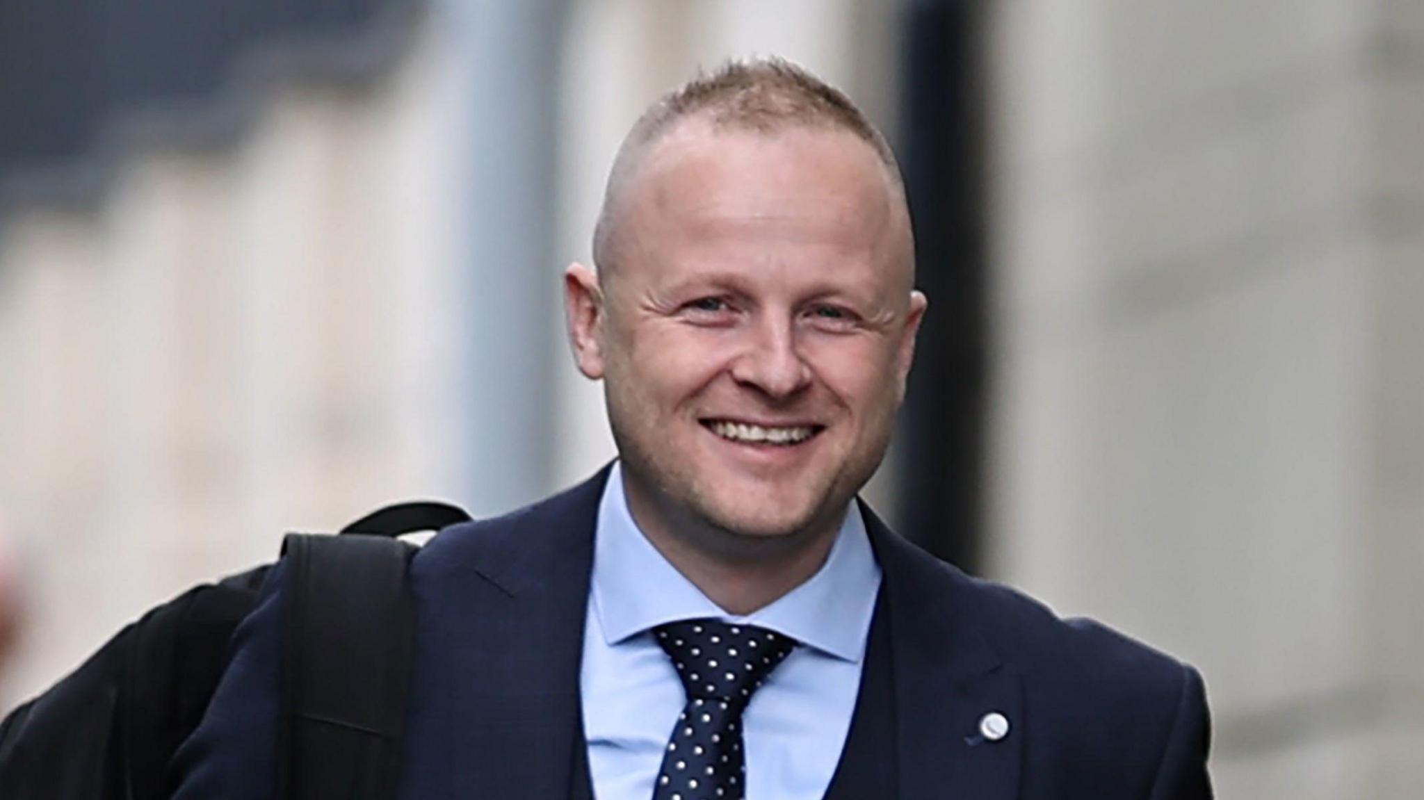 Jamie Bryson is wearing a dark suit and tie and is smiling as he walks to a court hearing. He has thinning fair hair and some stubble.