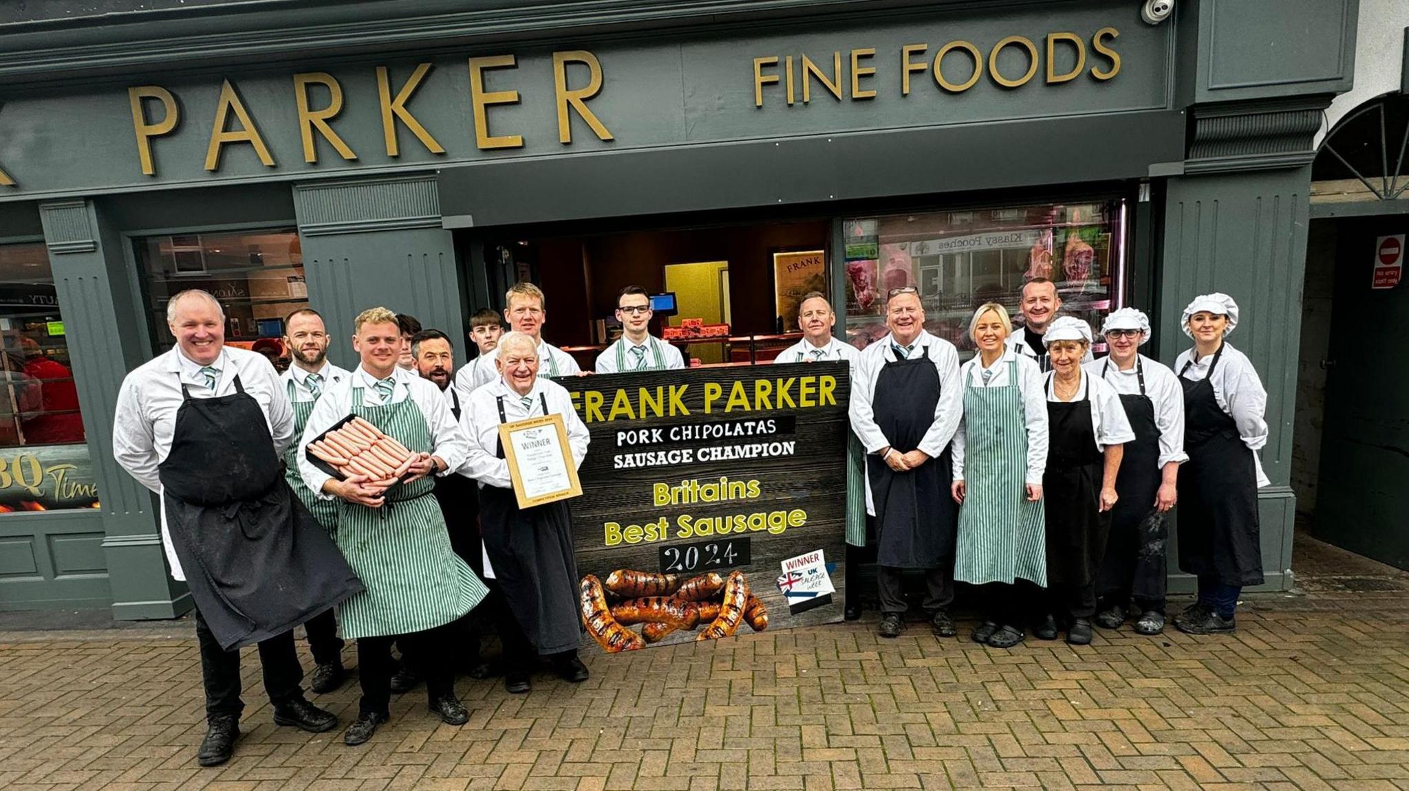 A group of men and women wearing aprons stand in front of shop. There is a sign in the middle which reads Britain's Best Sausage.