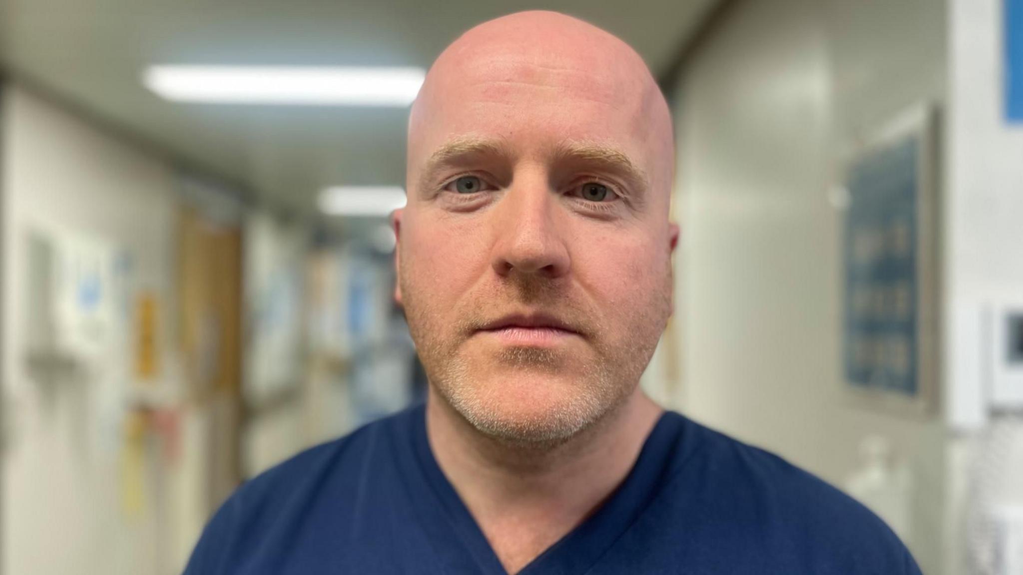 Head and shoulders photo of Dr McDonald looking directly at the camera. He is standing in a hospital ward but the background is blurred. He is bald-headed and has a blonde stubbly beard. He is wearing dark blue hospital scrubs.