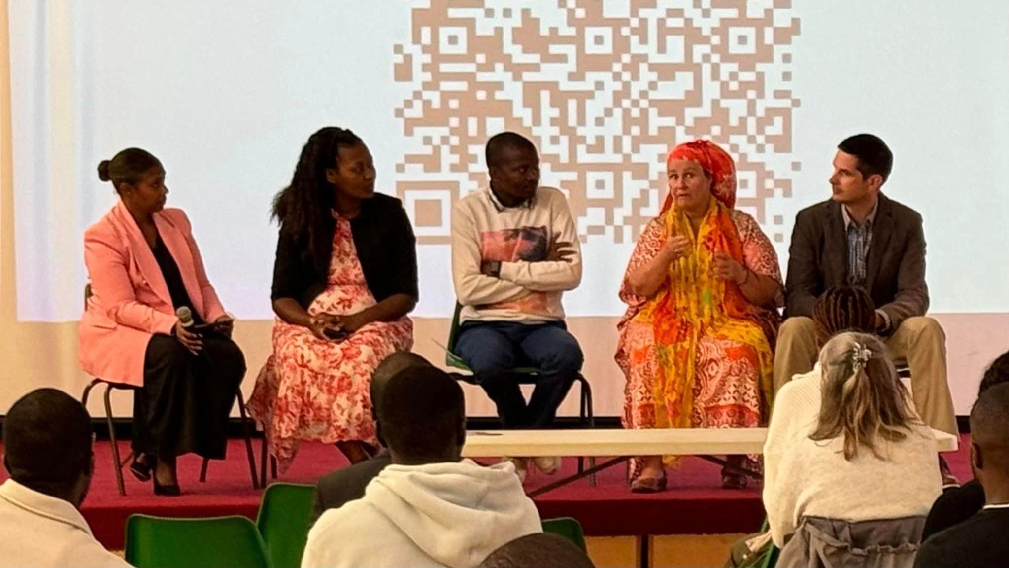 A panel sits on a stage as a woman in a bright dress speaks and the rest of the panel look at her. They are sat on a slightly raised platform in front of a group of people who are on plastic green chairs. Behind the panel, a projector screen shows a QR code.