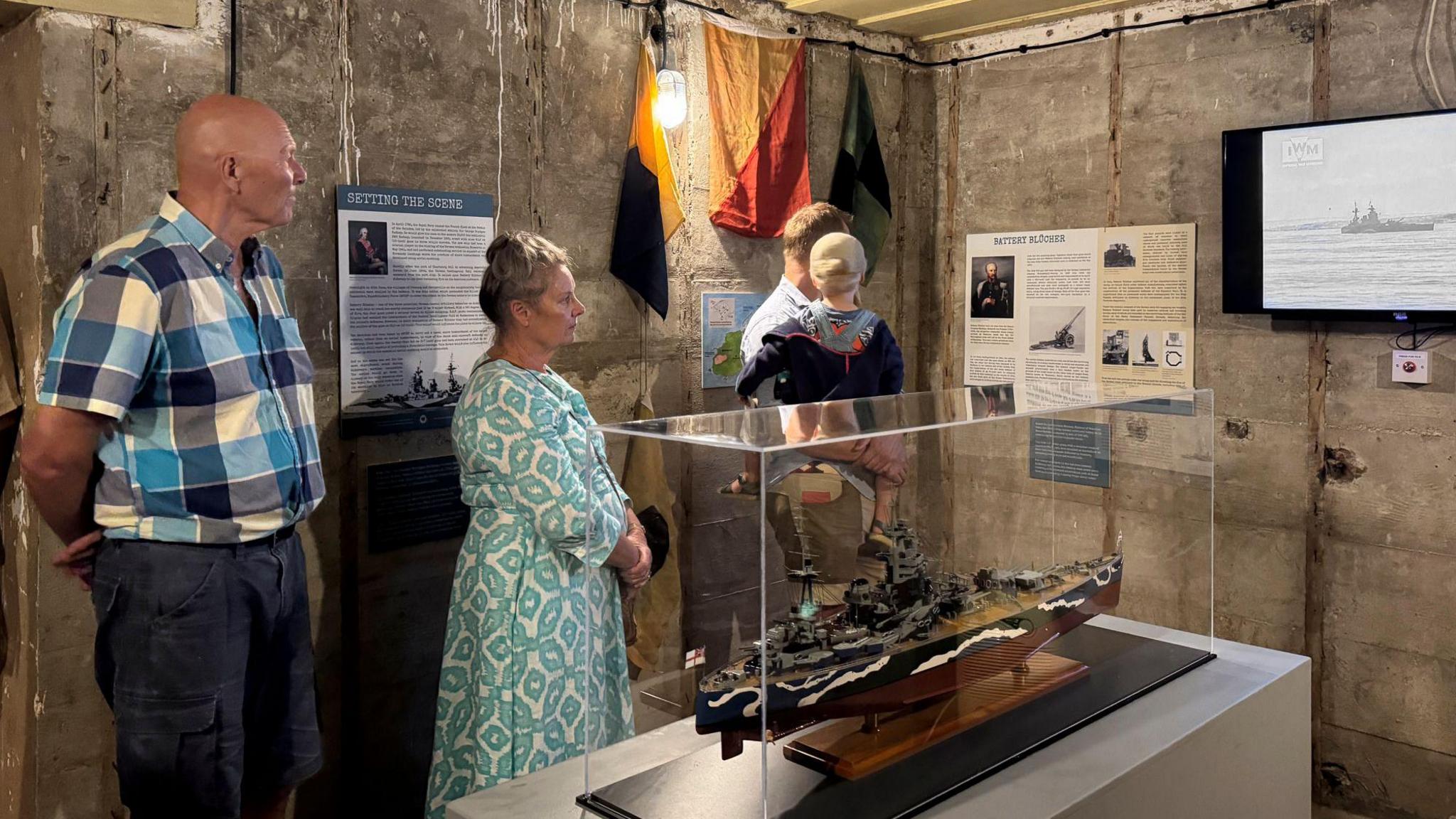 A man in a checkered shirt, a woman in a blue patterned long sleeve dress, a man wearing a blue shirt holding a small child with a hat on and a navy coat all standing in a bunker with signs on the walls and a small scale version of a navy ship