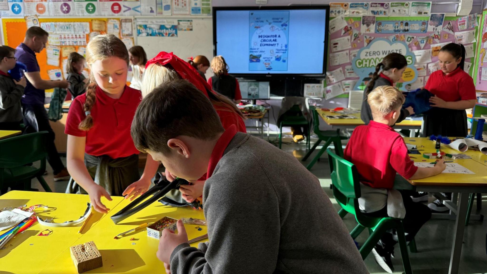 Pupils are hard at work as they begin creating their crafts. Some are stood up beside their yellow desks, while one boy on the right hand side of the picture is sat down using a pencil and paper.