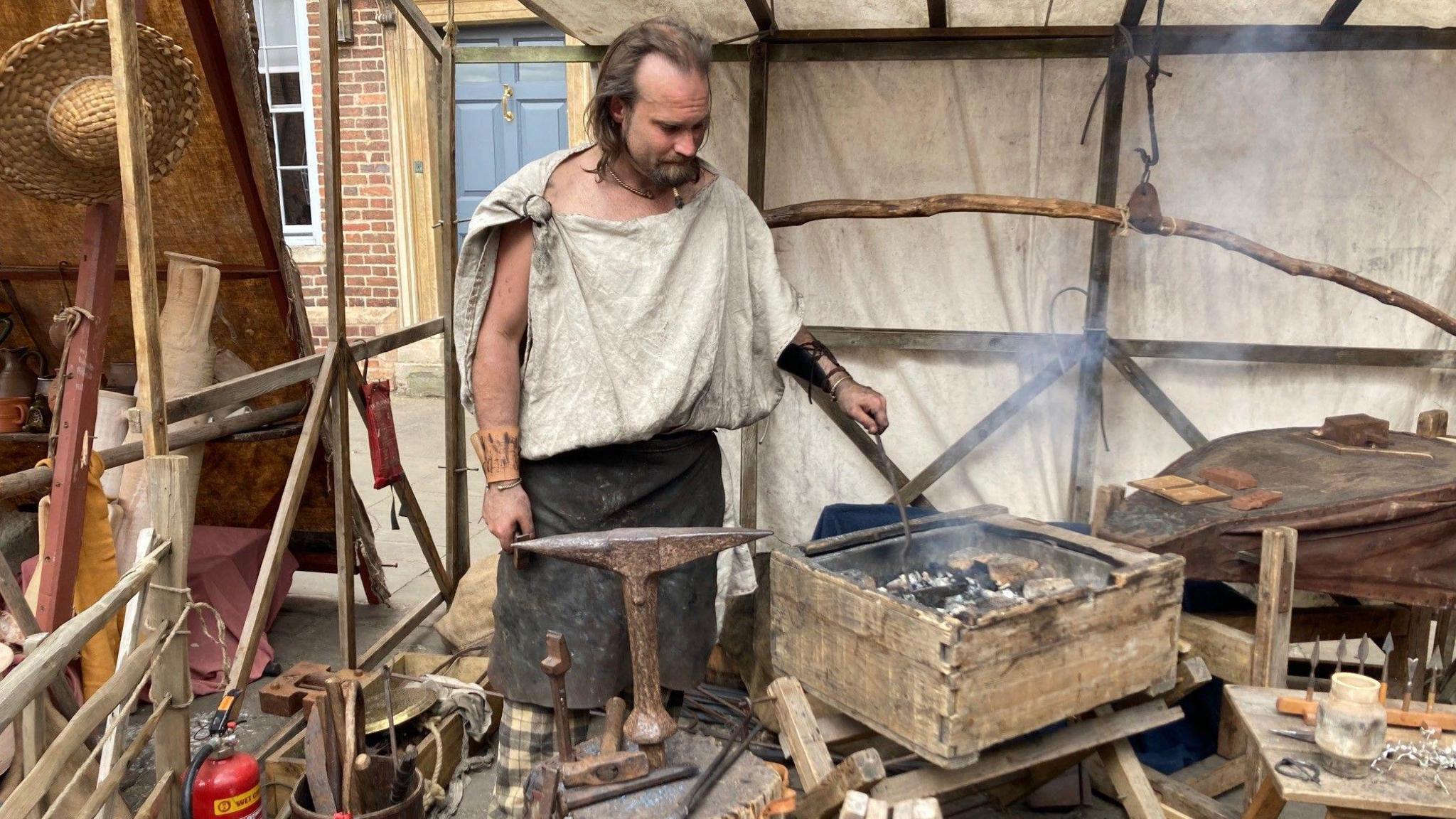 A man stokes a fire as he demonstrates how a Roman blacksmith would have worked.  He wears an old apron and a toga-style top tied at the shoulder.