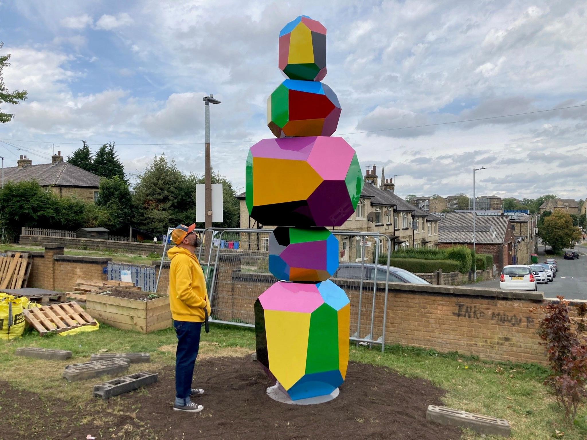A man in a yellow hooded top and baseball cap stood looking up at a 4m multi-coloured sculpture with five separate multi-sided sections.