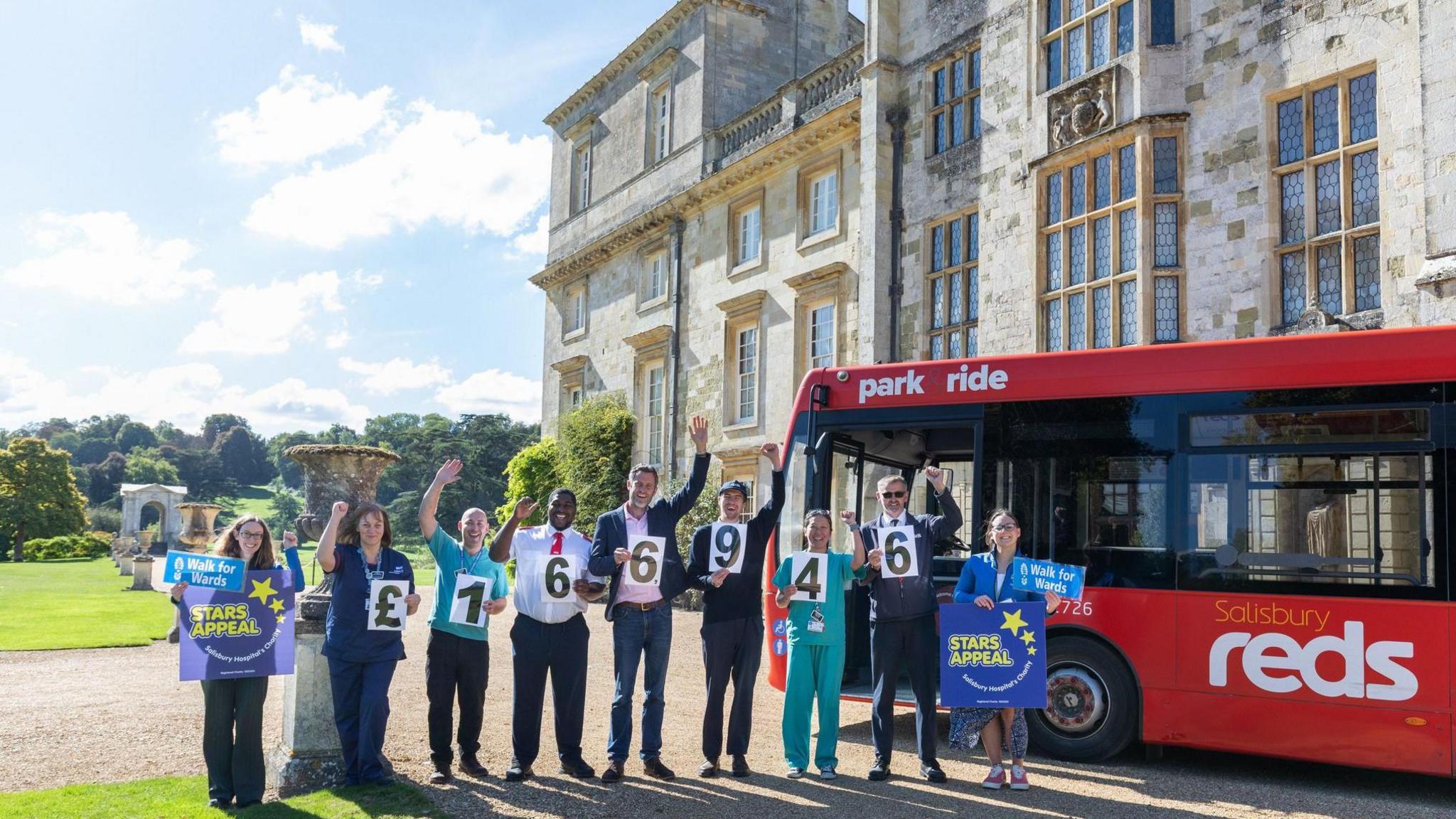 People stand in a line on a sunny day in the gardens outside a large stately home. They are all smiling and waving at the camera, each holding a sign. on either end, people hold the logo for the charity and in the middle each person holds a number to spell out £166, 946. To the right, a red bus is parked, the company being one of the sponsors.