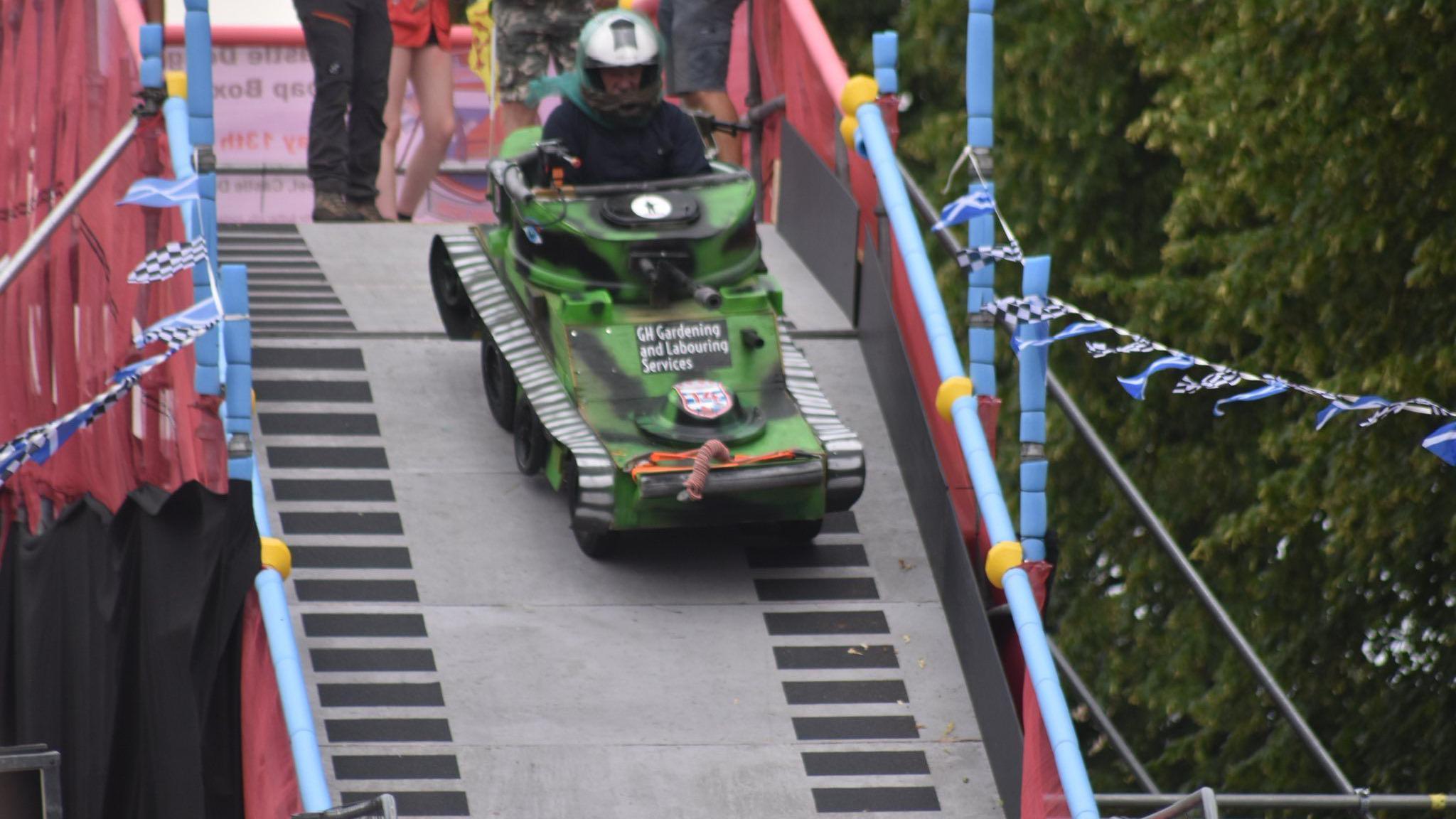 A close up of a small green and black camoflague tank rolling down the ramp. At either side there are checkered and Scotland flags.