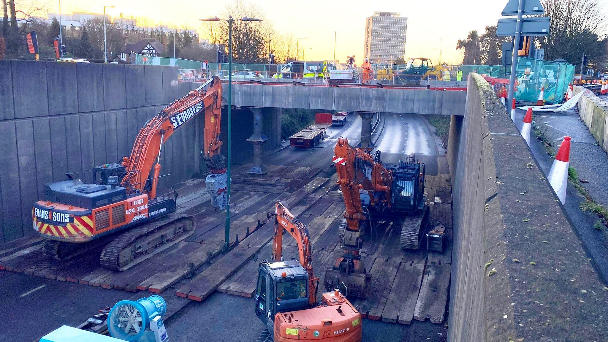 A scene of intense roadworks activity on overpass bridge with a dual carriageway underneath and three construction machines. 