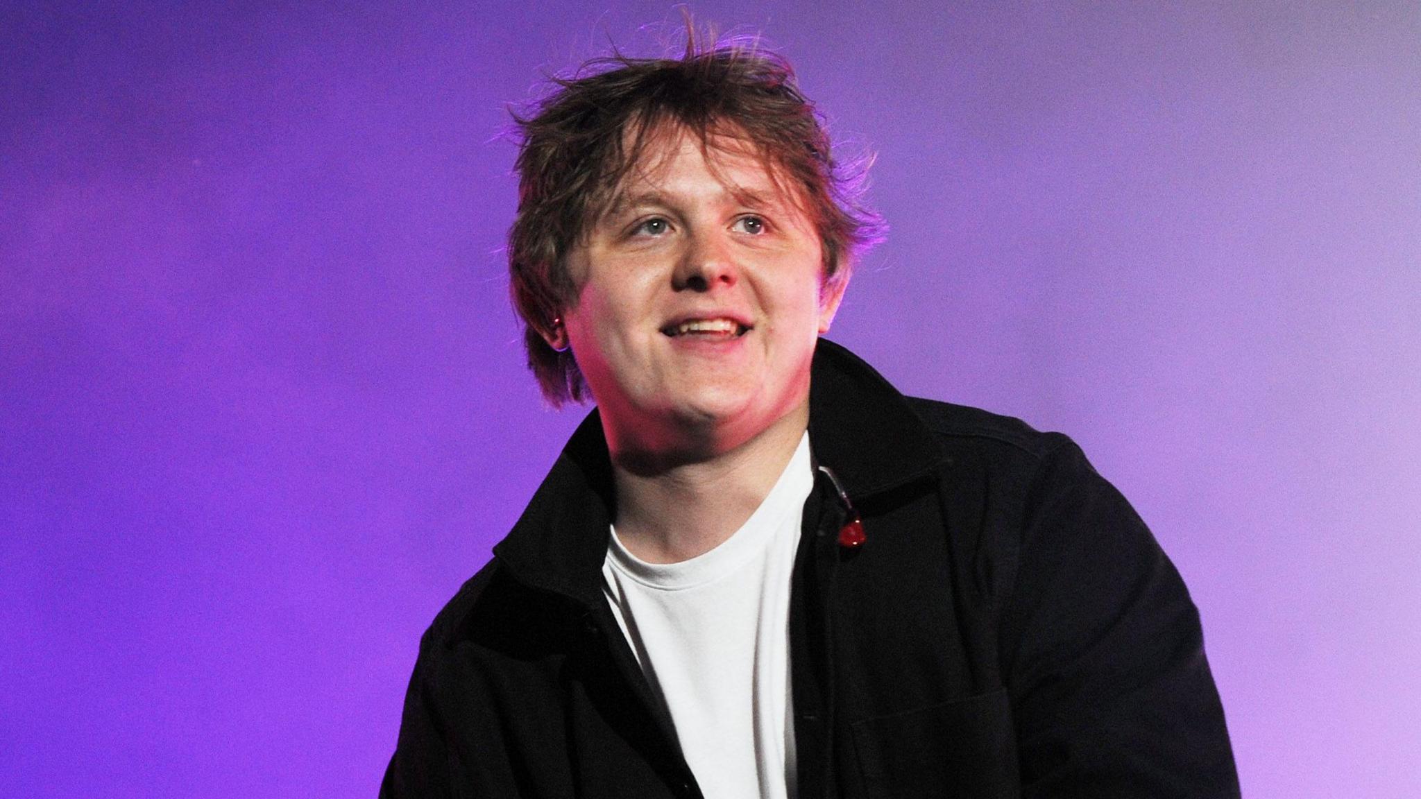 Lewis Capaldi performs onstage - he is wearing a white shirt and black jacket, is holding a microphone and smiling