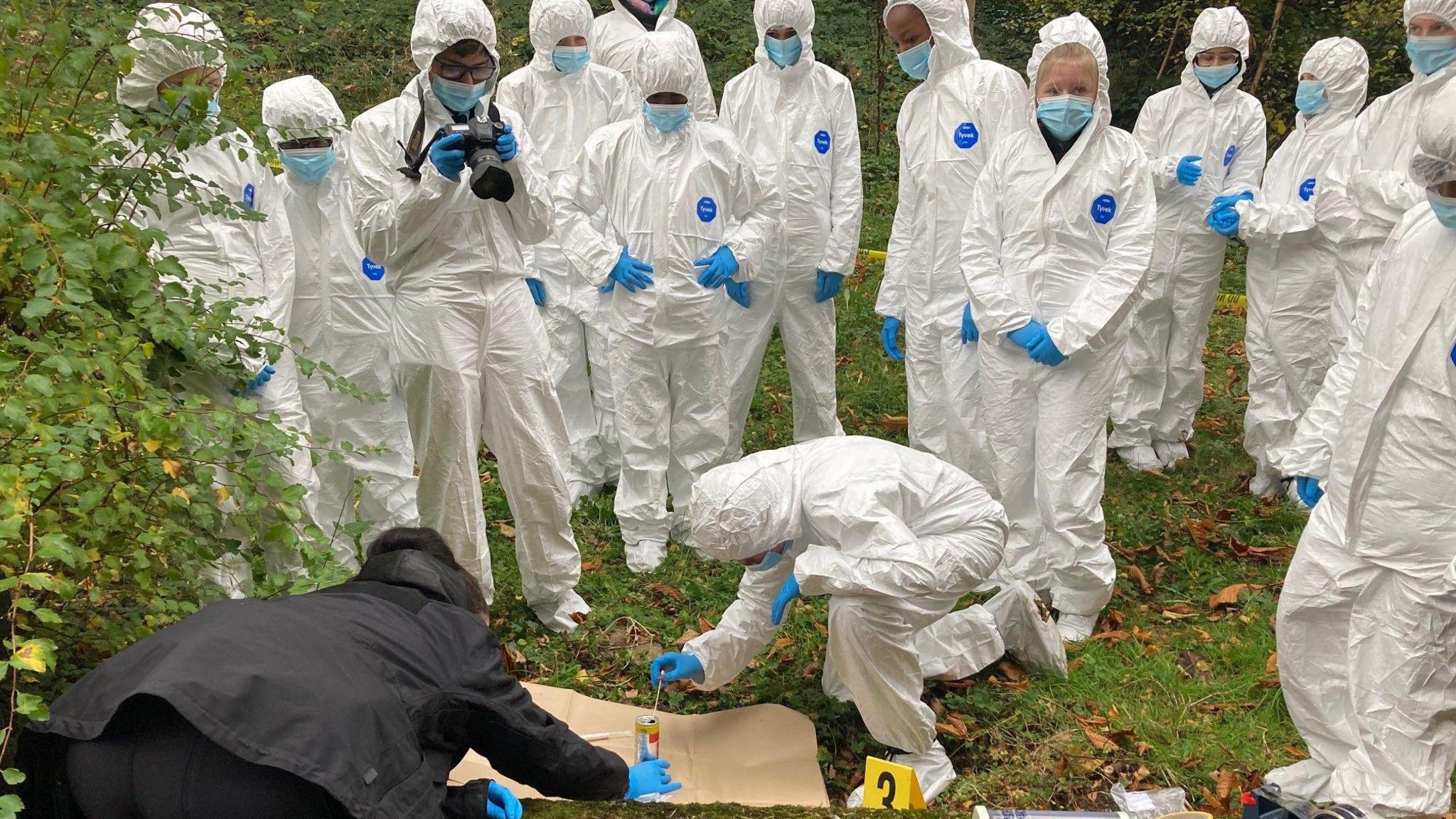 School children wearing white protective overalls and blue rubber gloves inspect a mock-up of a crime scene with the help of a police officer who is wearing a black coat and trousers.