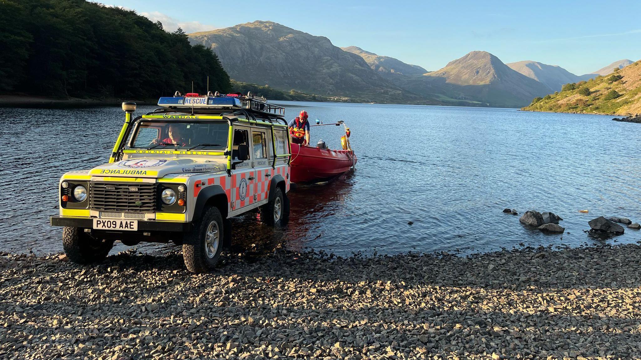 A mountain rescue Land Rover backing into the water on a pebble beach. A red boat is attached to the back of it. Mountains can be seen in the background.