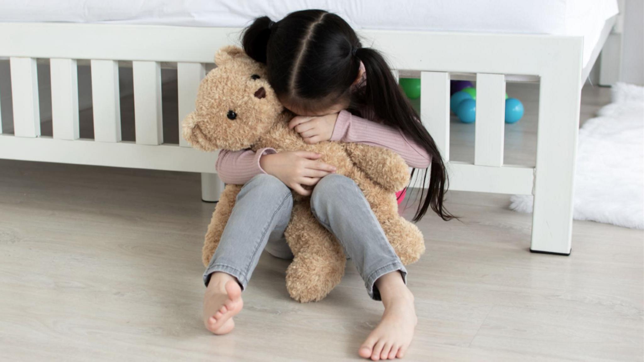 A girl with her hair in bunches sits on the floor in front of a bed. She seems to be crying but her face is hidden by the teddy bear she is holding in her arms. 