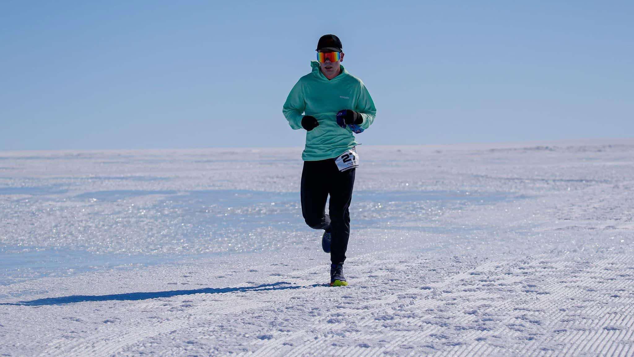 Paul running in Antarctica on the snow. He's got his colourful glasses on with a light blue hoodie and long, black trousers.