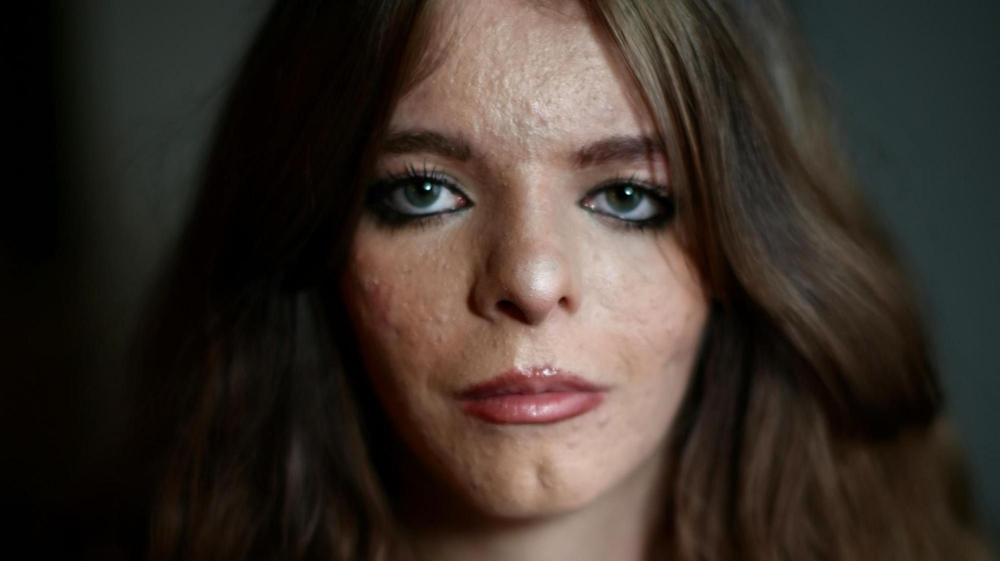 Amy looks at the camera. She has thick dark brown hair which is parted in the middle and is wavy. Her eyes are green. She is wearing makeup with gloss lipstick on and eye liner with mascara and brown brow liner. The light is caught in the pupils of her eyes and she is in front of a dark background. You can see her face and neck.