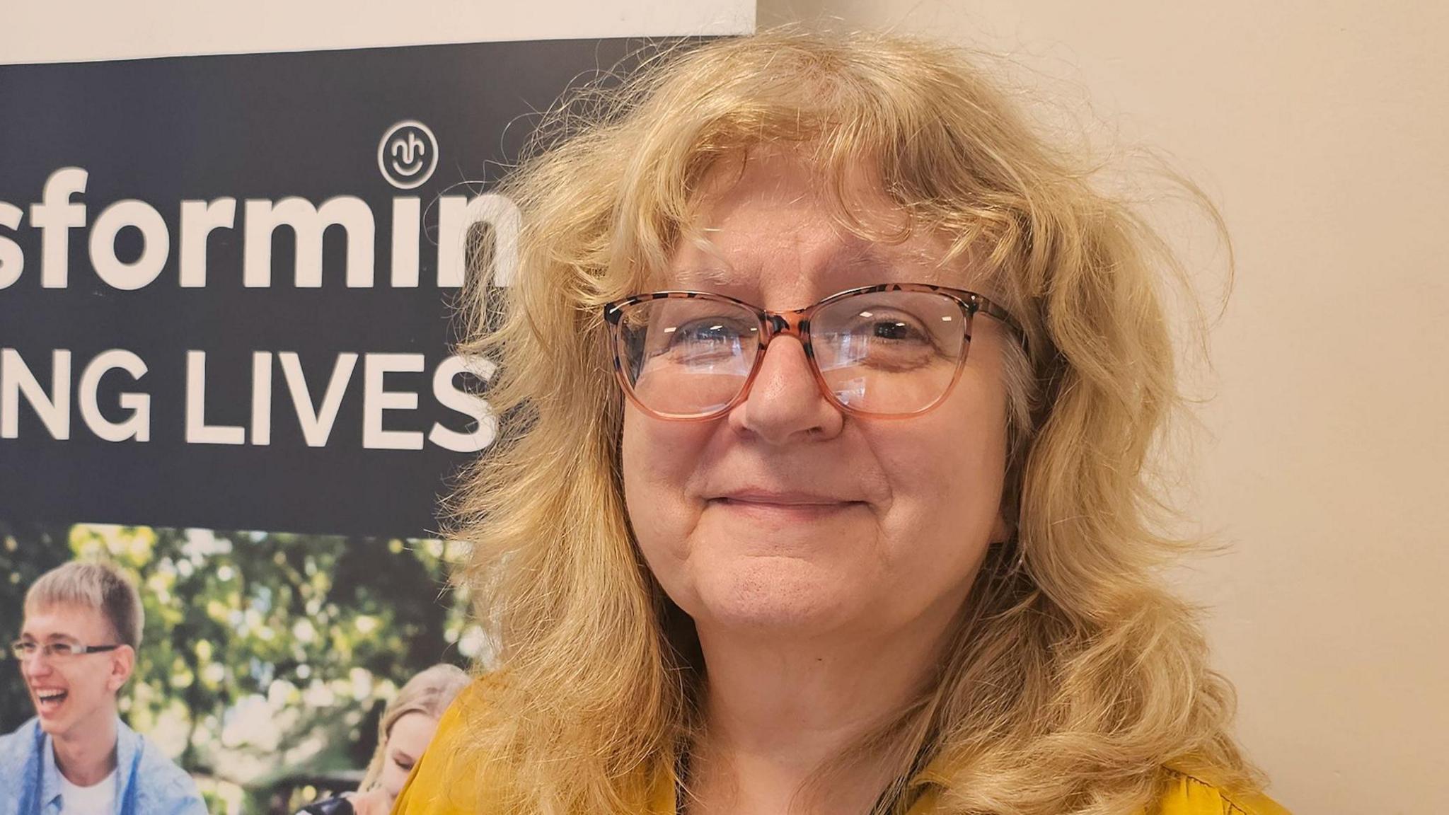 A woman with curly blonde hair and tortoiseshell glasses, smiling at the camera. She is wearing a yellow top.