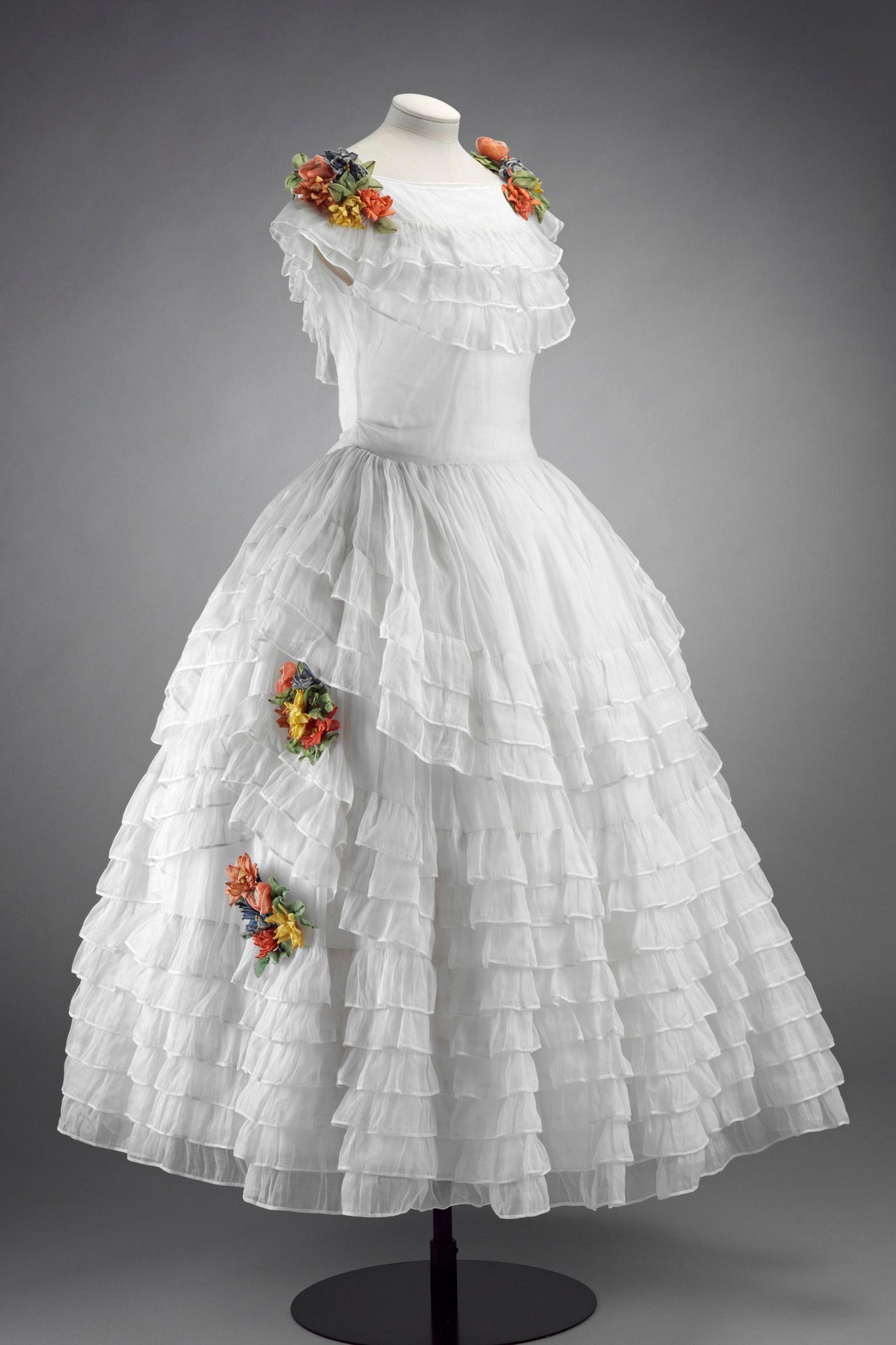 A white 18th-century-style dress with multiple tiers of sheer ruffles and clusters of colourful artificial flowers on the shoulders and skirt, displayed on a mannequin.