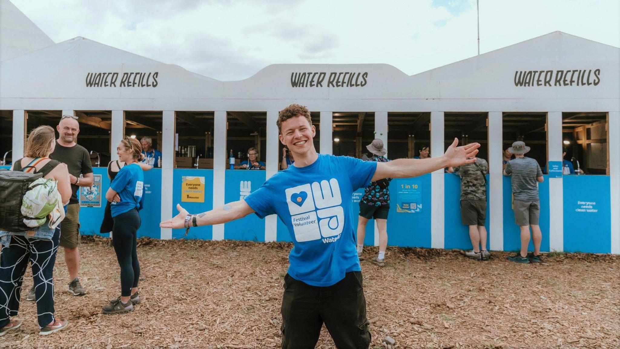 Conor Blundell wearing a blue wateraid t-shirt, standing with his arms outstretched and smiling in front of a water refills station at Glastonbury