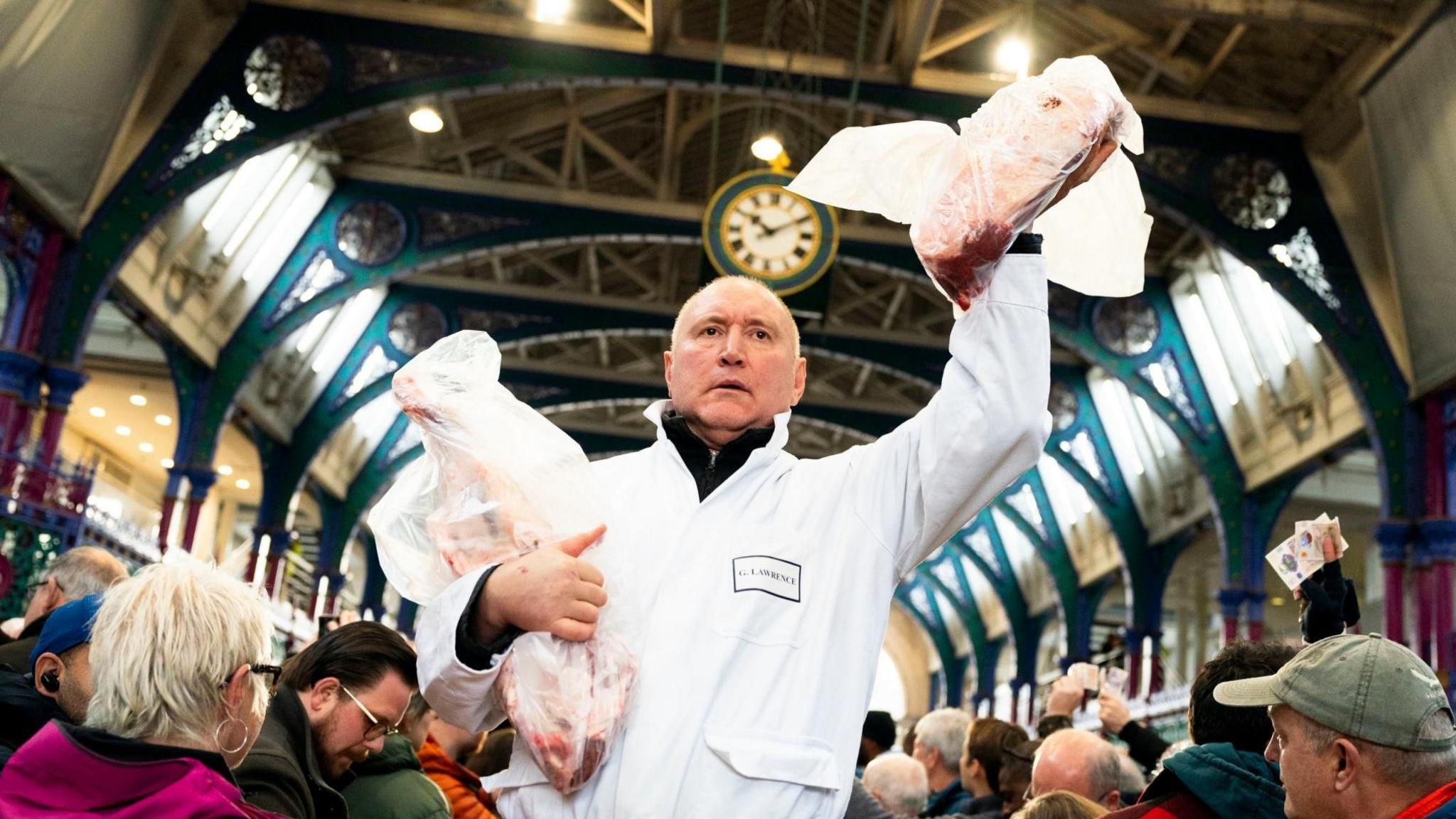 A trader wearing a white overall holds to joints of meat - one of which in the air - while people stand around him in Smithfield meat market