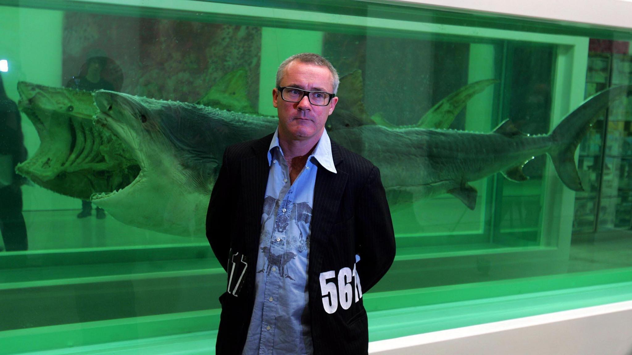 A man in a baggy blue shirt and black jacket with numbers on the side stands in front of a piece of artwork made of a shark, cut lengthways and suspended in formaldehyde.