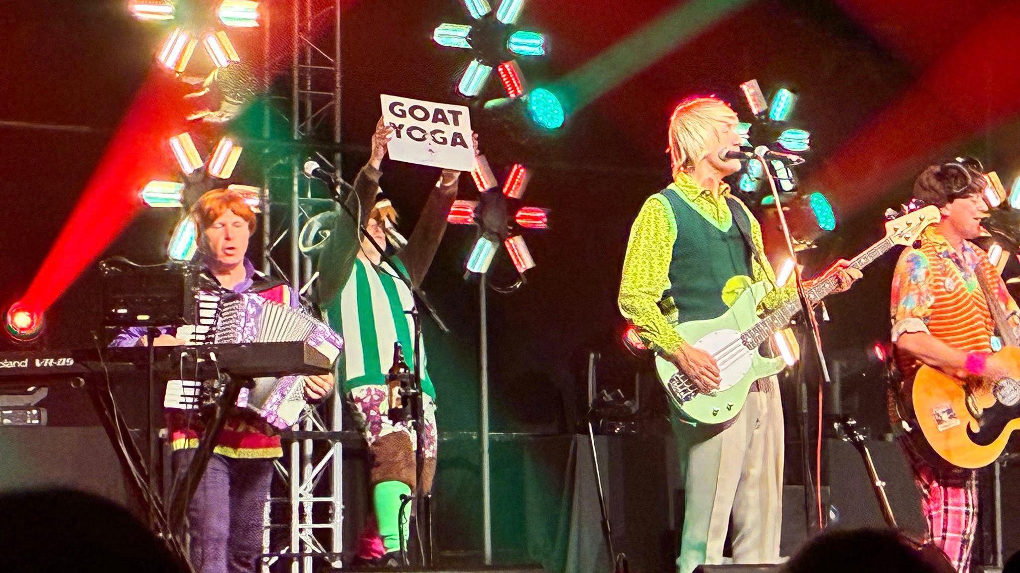 Four men playing musical instruments on a stage that is lit by red and blue lights. They are wearing brightly coloured outfits and have short wigs on. One is playing accordion, another is holding a sign that says "Goat Yoga", while there is a bass player and guitarist alongside him.