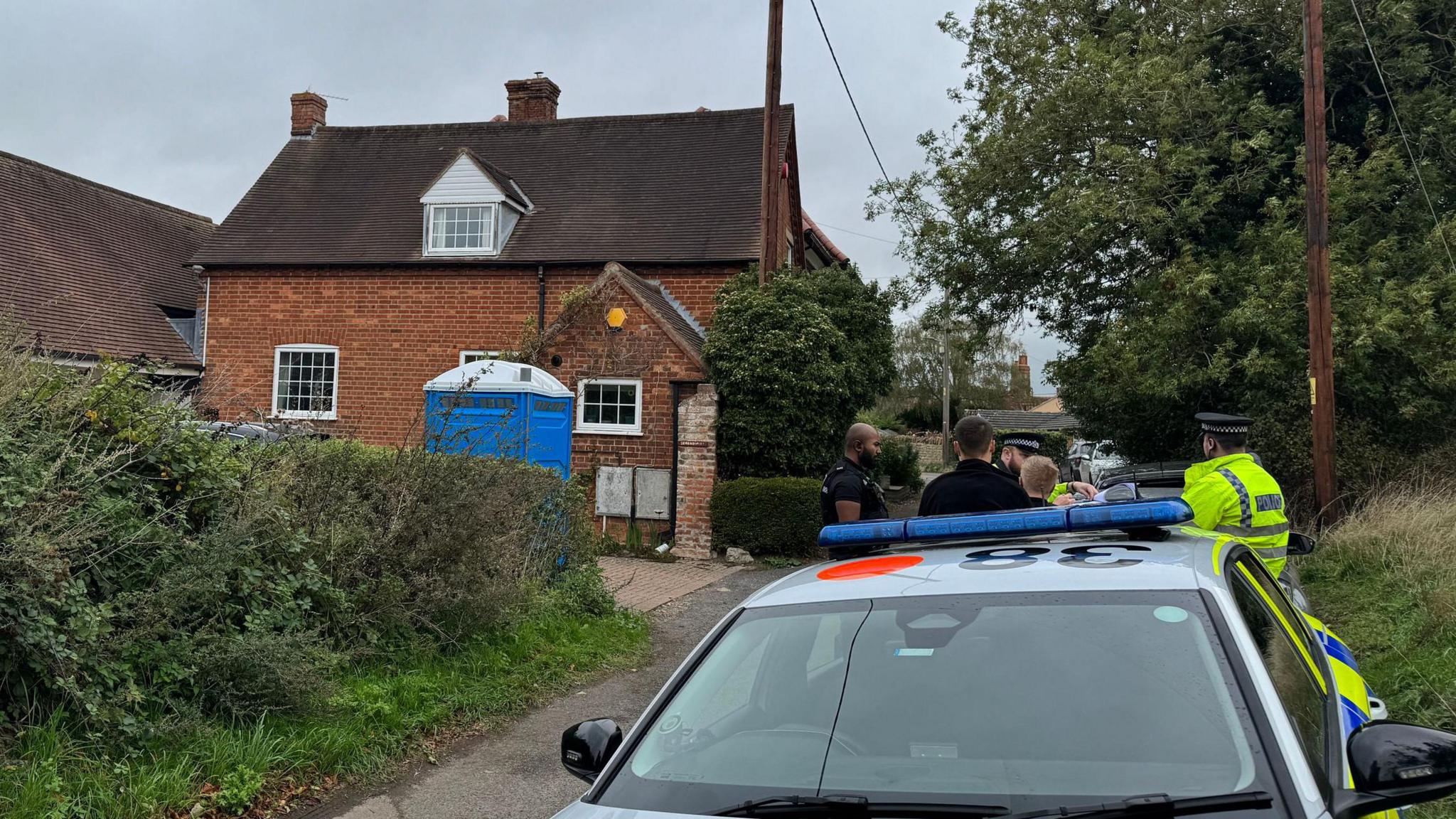 A group of police officer, mostly in hi-vis jackets, are crowded around opposite a large detached red brick house.