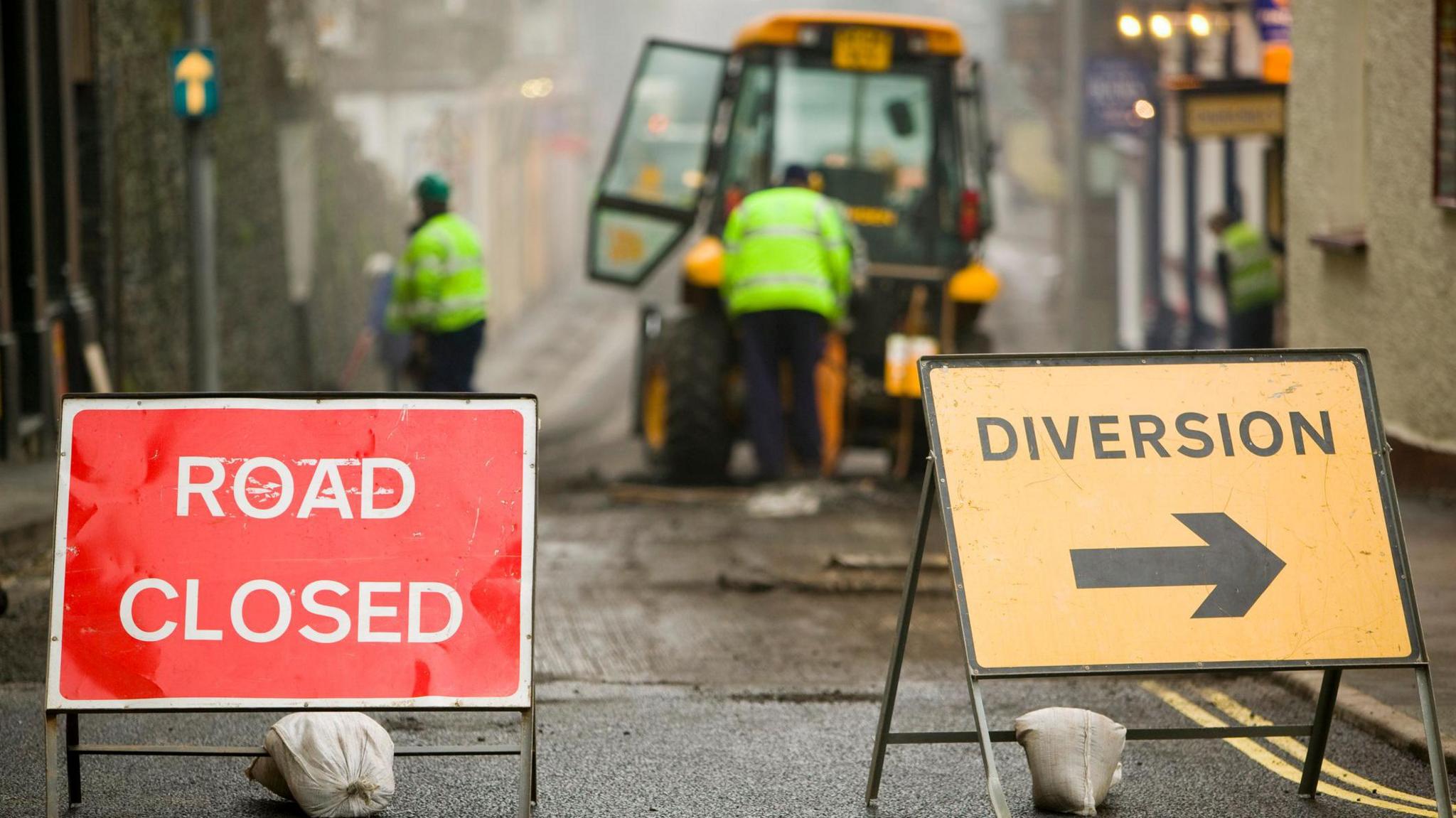 A red sign that has 'road closed' in white letters and a yellow sign with 'diversion' in black letters stand in the middle of a road. Behind the signs there are men in high vis jackets working on a road and a yellow tractor.