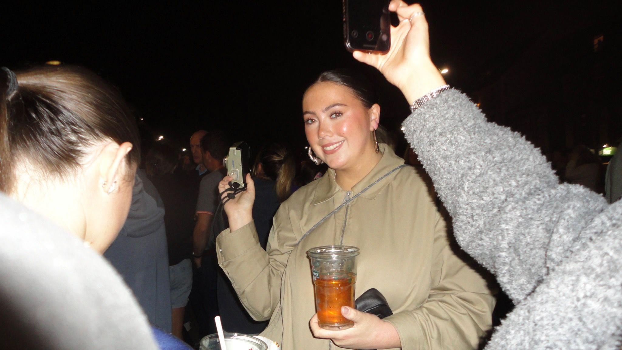 Aoibheann in a beige jacket is at a crowded pub, holding a drink and a small camera, with others around holding phones.